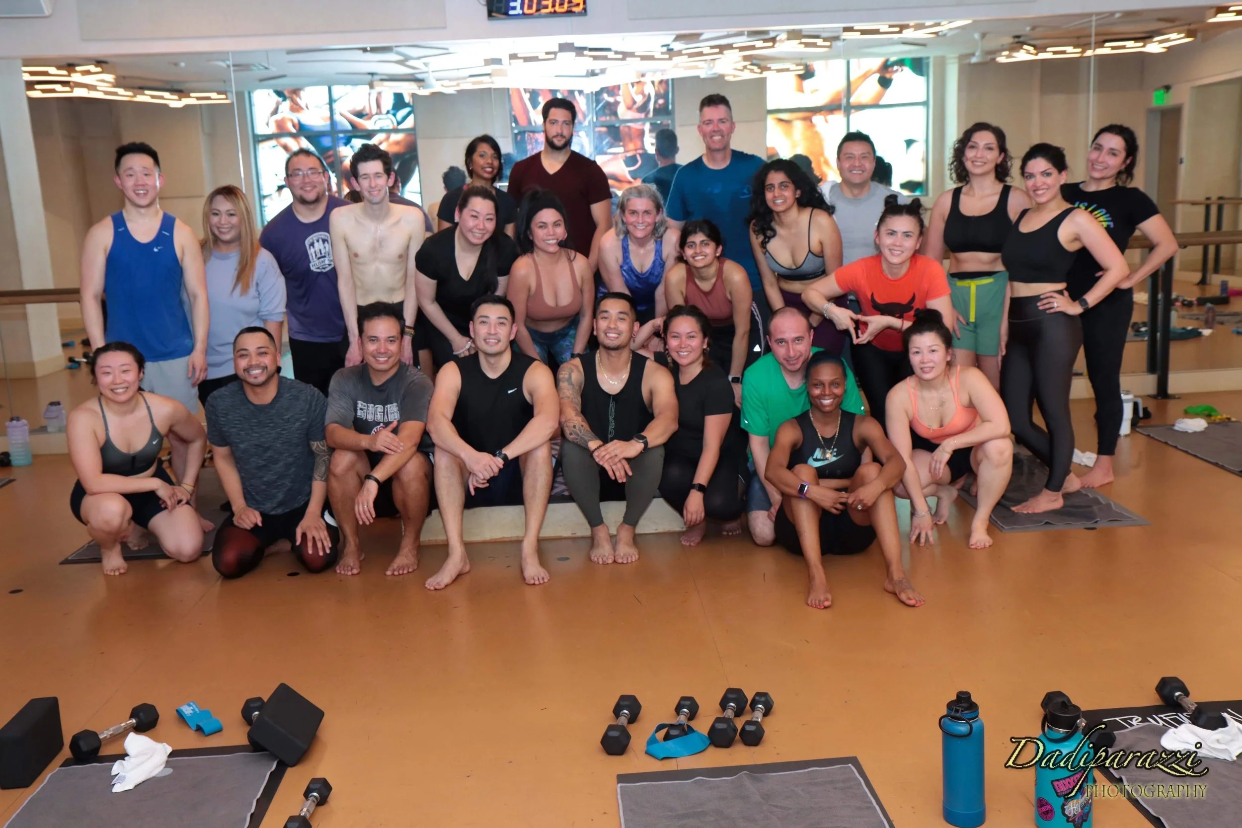 Group of people in a fitness class, smiling and posing indoors with workout equipment visible in front.