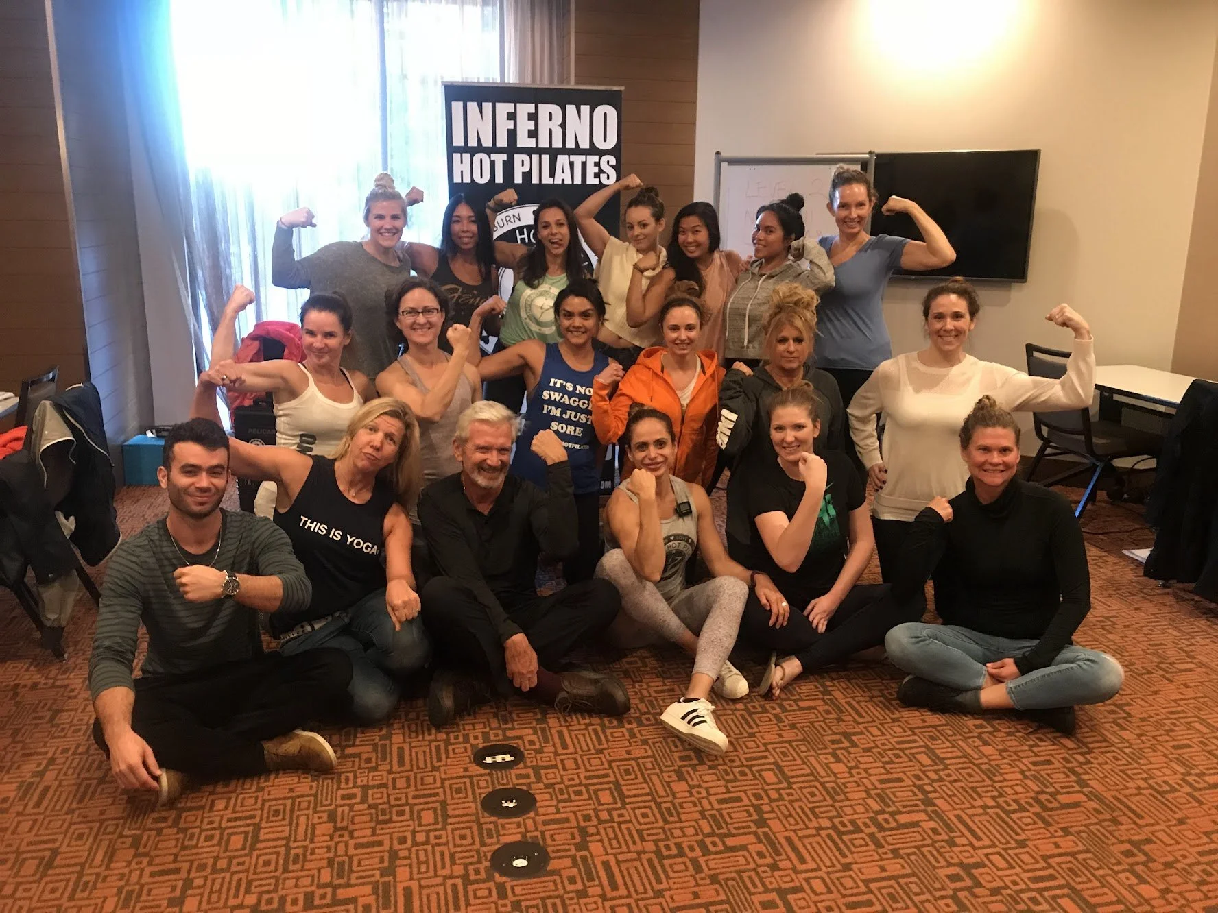 A group of women and men in workout clothes posing together in a fitness class or workshop, flexing their biceps.