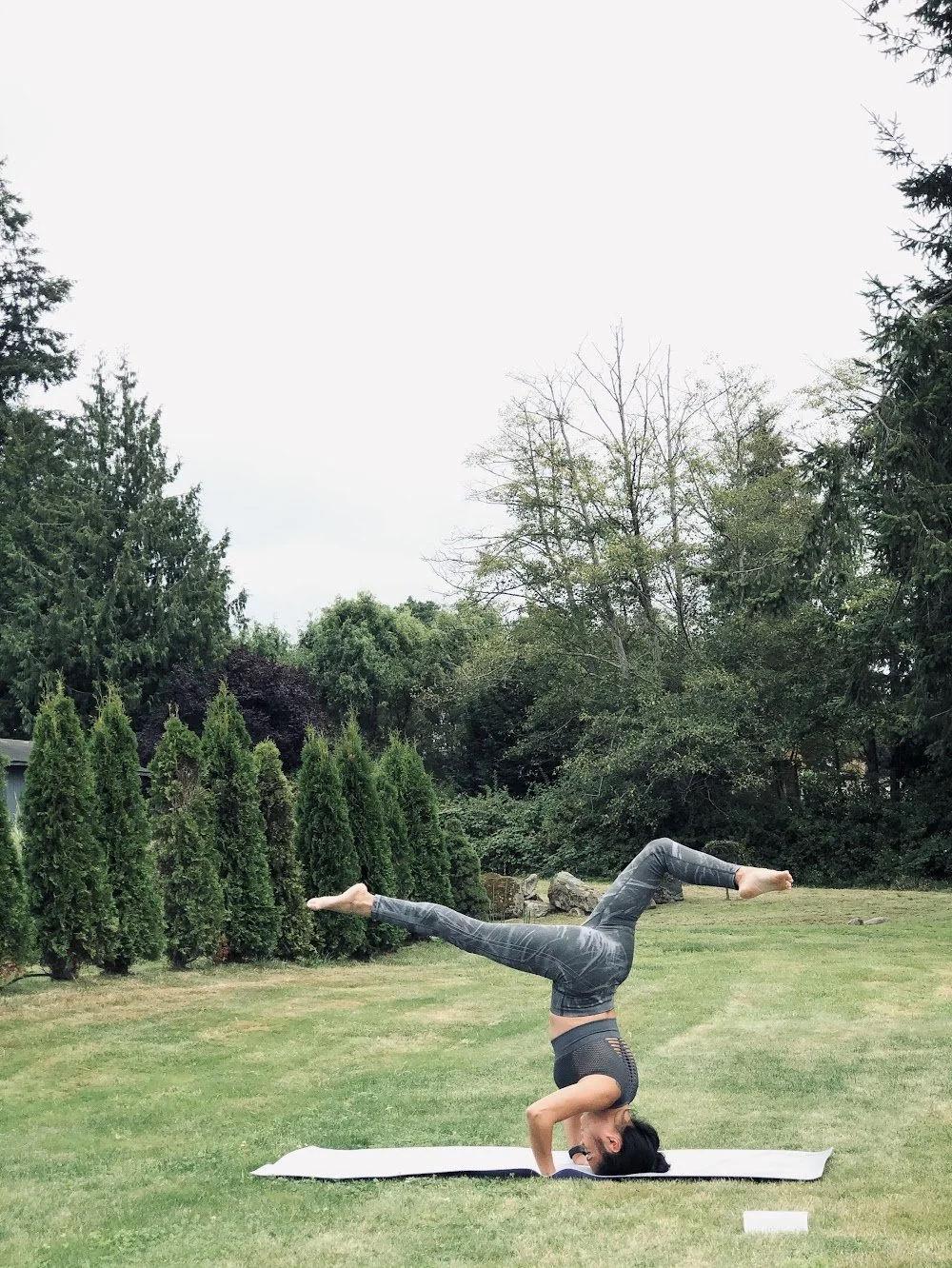 A woman practicing yoga outdoors on a grassy lawn, balancing on her head with her legs extended in a split position, on a yoga mat surrounded by trees and bushes.