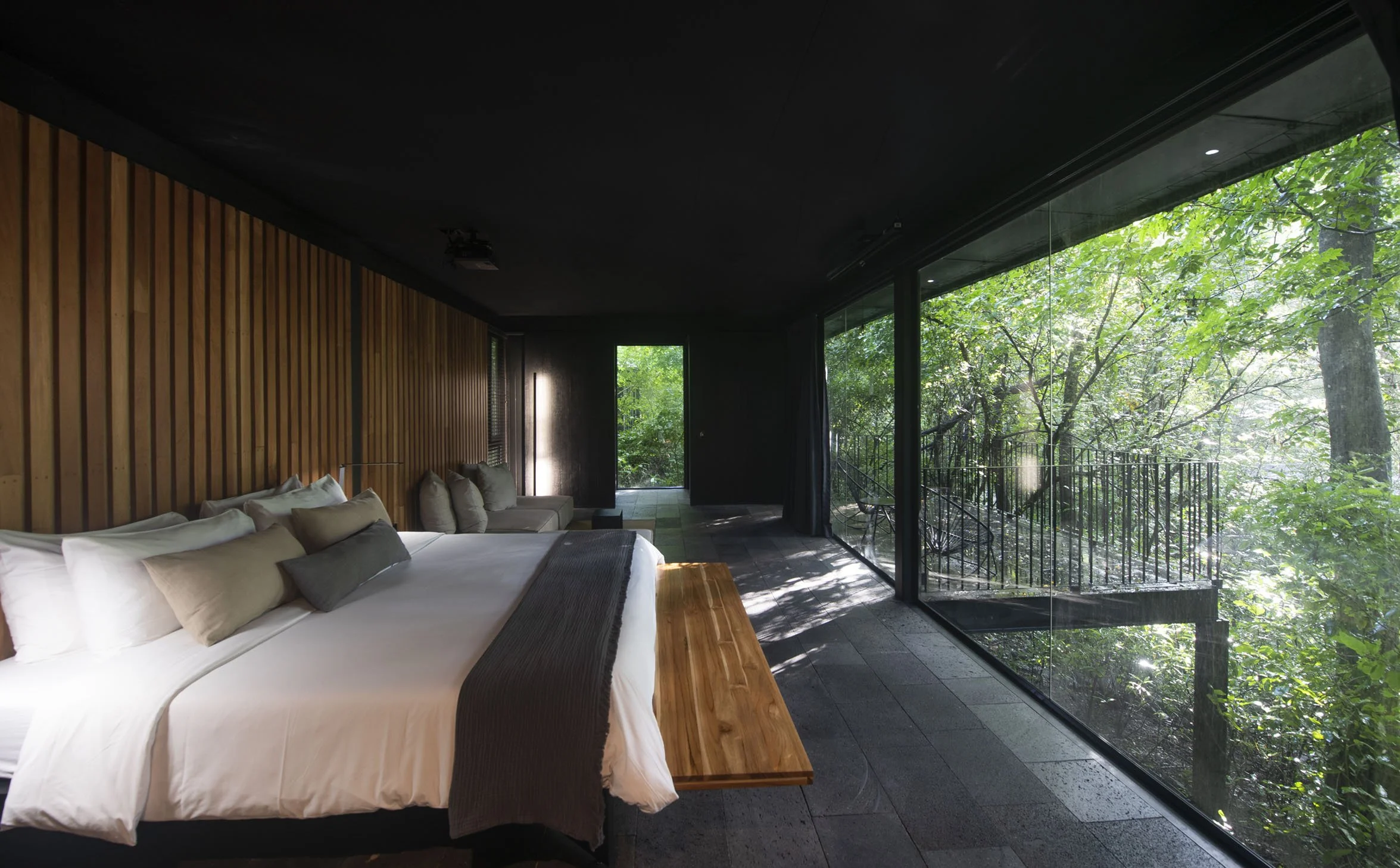 Modern bedroom with a large bed, multiple pillows, and a wooden bench, featuring floor-to-ceiling windows overlooking a lush green forest.