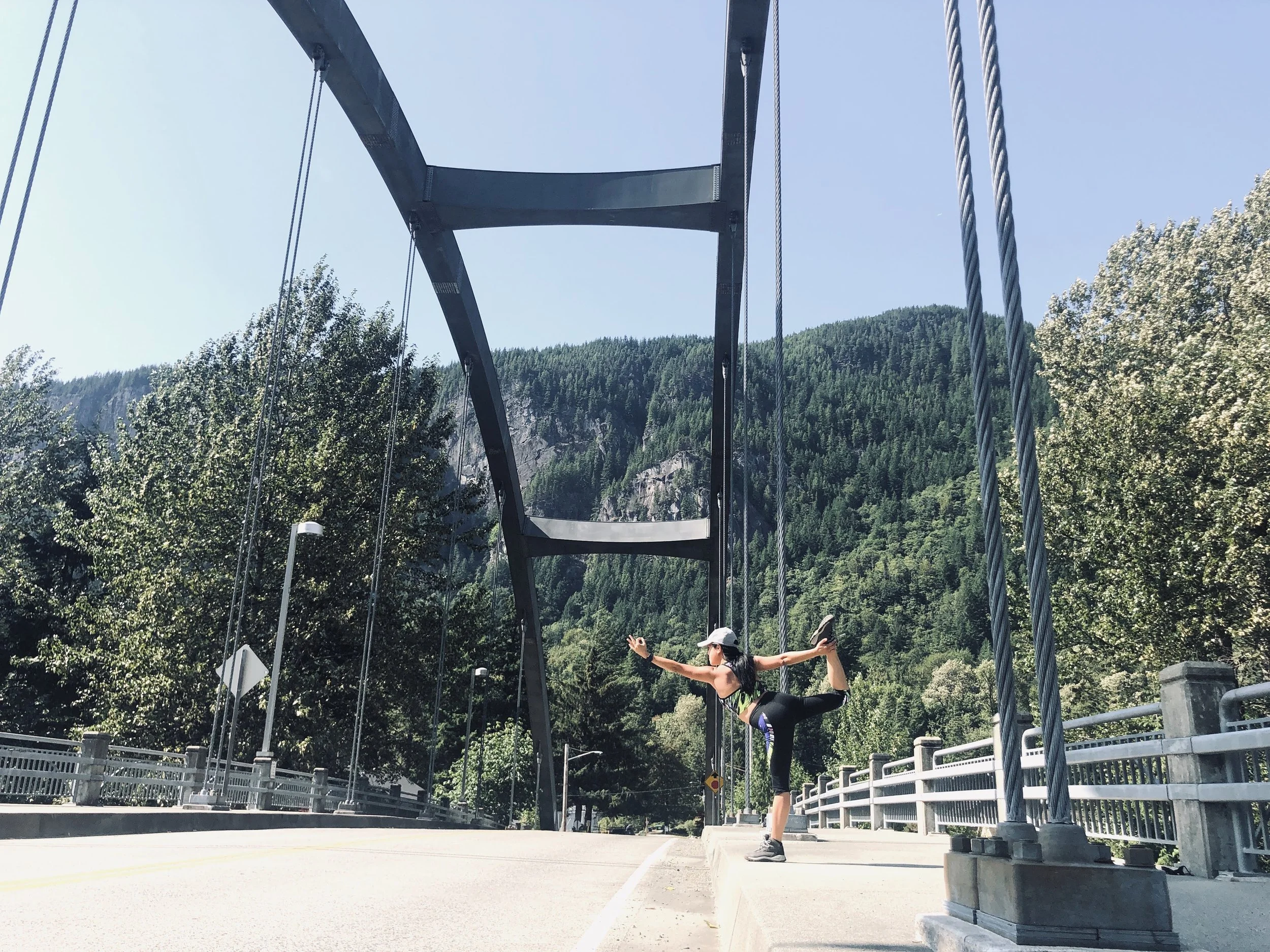 A woman in athletic clothing doing a yoga pose on a sidewalk in front of a suspension bridge with a mountain and forest in the background.