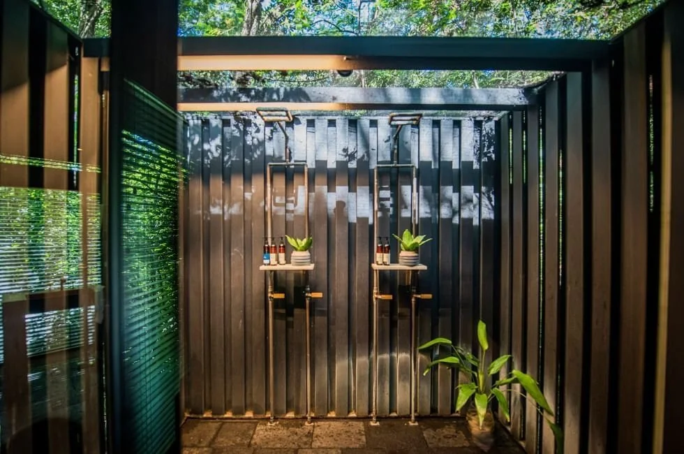 An outdoor shower with dark wooden slat walls and ceiling, two showerheads mounted on the wall, and small shelves holding potted plants and bottles, surrounded by green foliage.