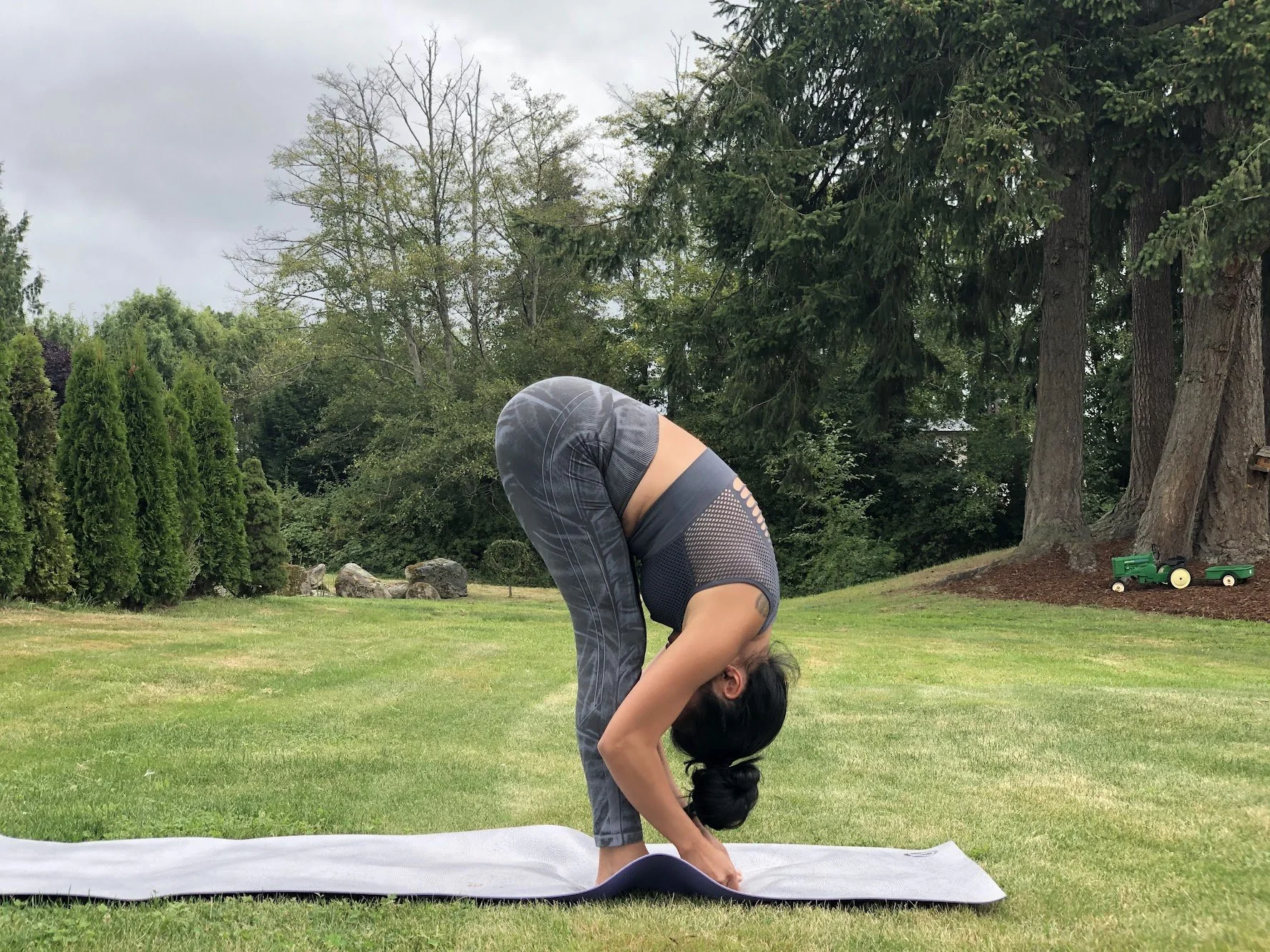 A woman practicing yoga outdoors on a grass lawn, positioned in a forward bend pose with hands touching the ground and head down, with trees and greenery in the background.