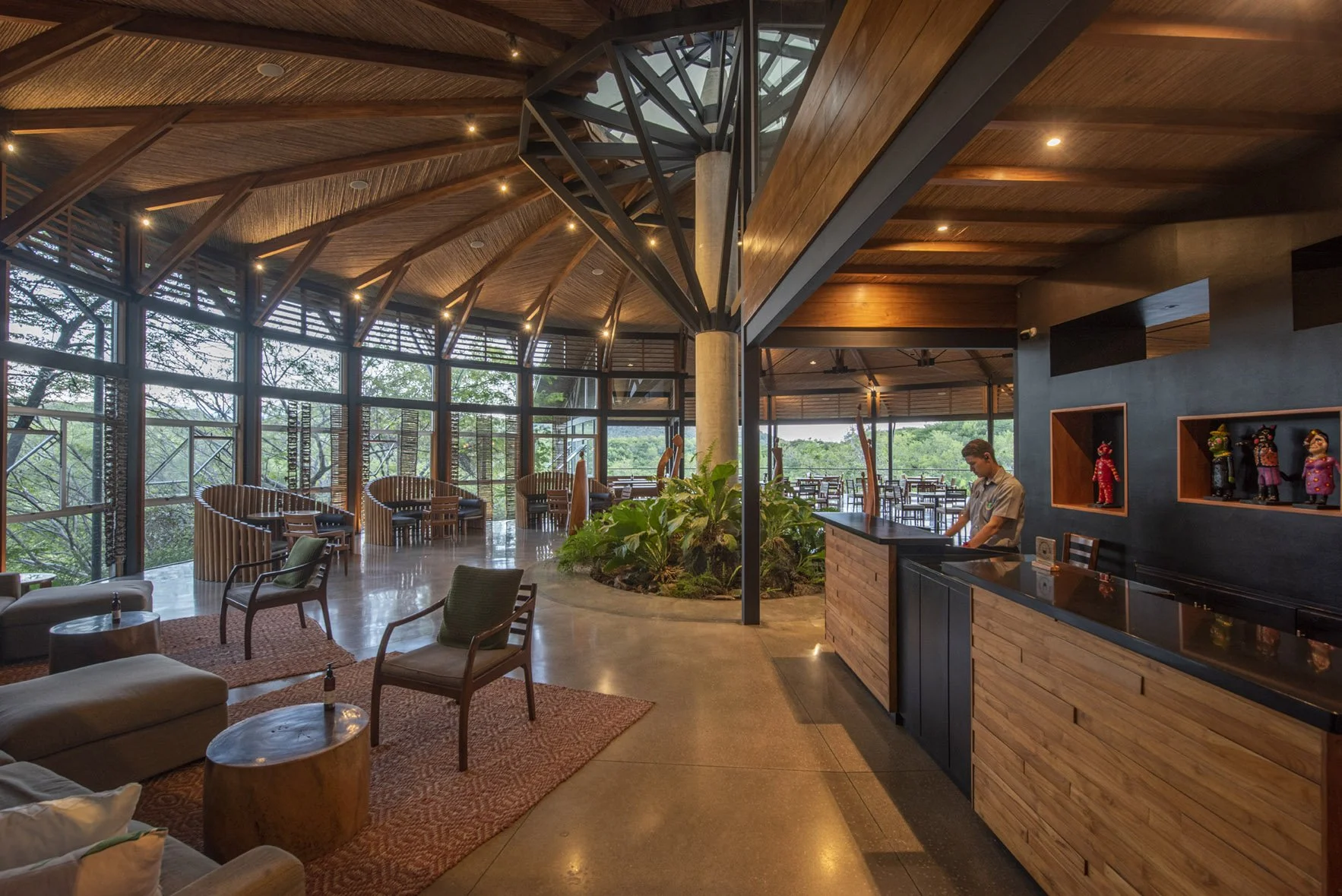 Interior of a modern restaurant or cafe with large glass windows, wooden ceiling, lounge chairs, a small bar area with a person working behind it, decorative figurines on shelves, and outdoor seating visible through the windows.