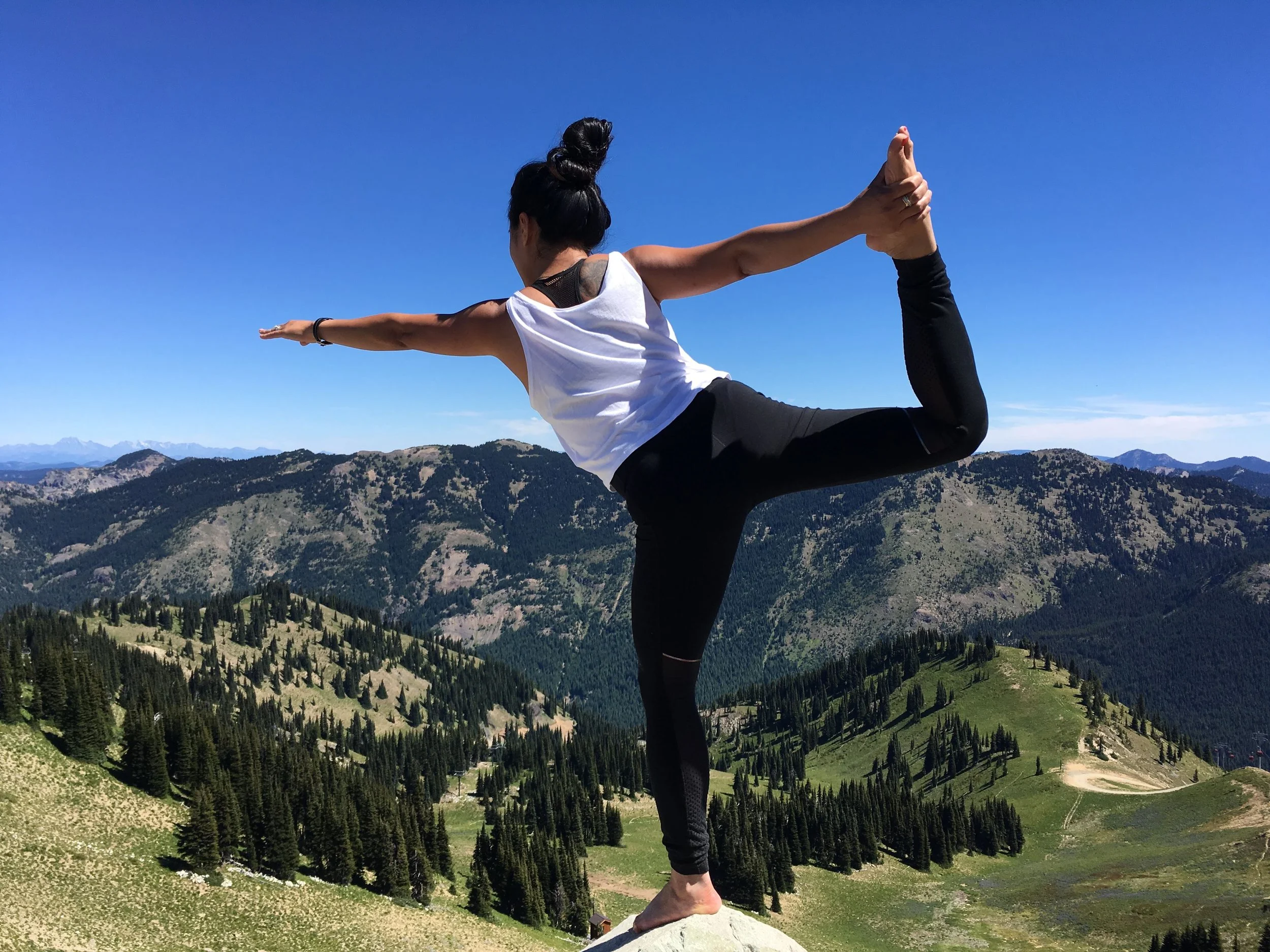 A woman practicing yoga on a rock in a mountainous landscape with pine trees and a blue sky.