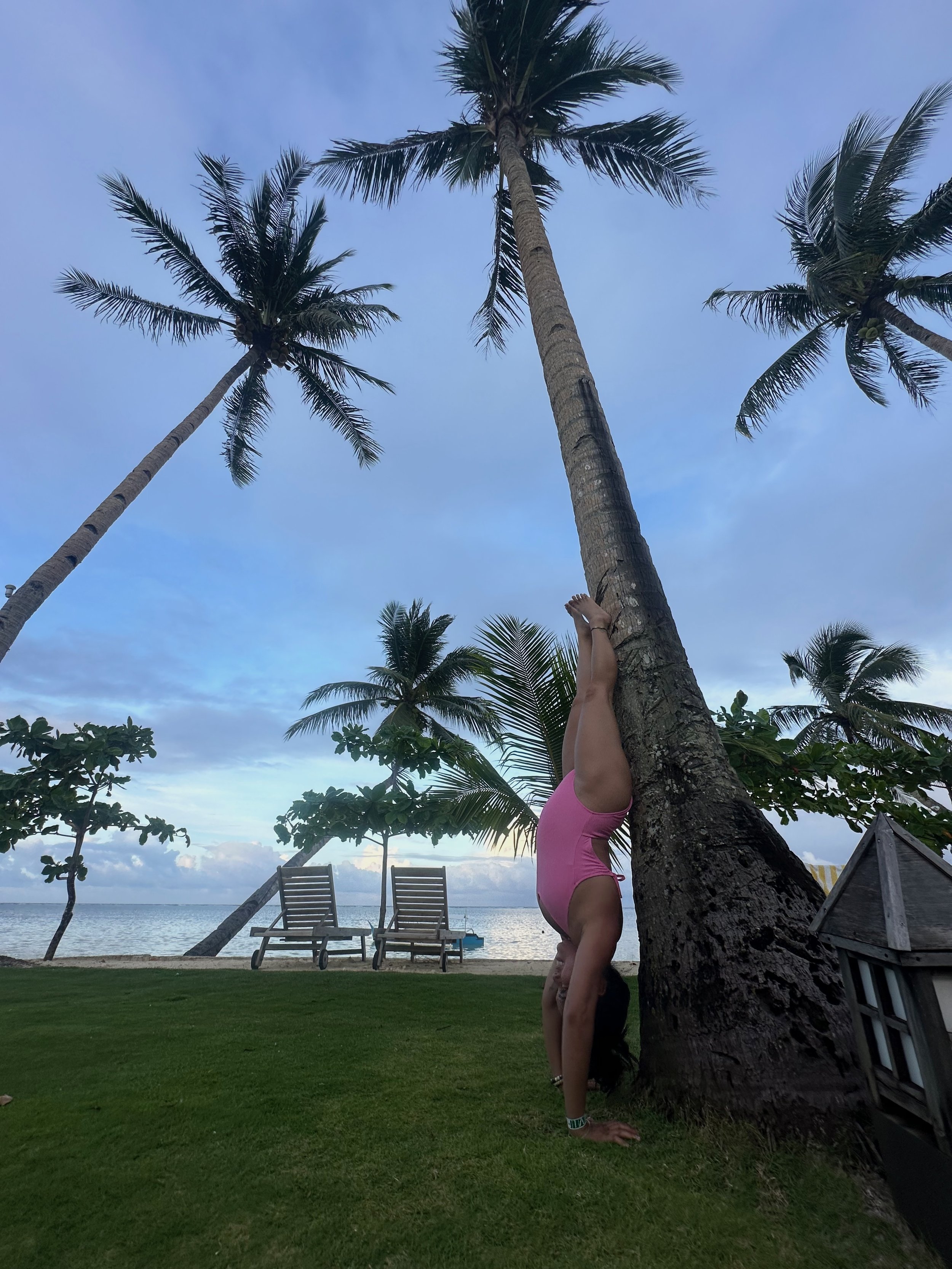 A woman in a pink swimsuit performing a handstand against a palm tree near the beach with lounge chairs and the ocean in the background.