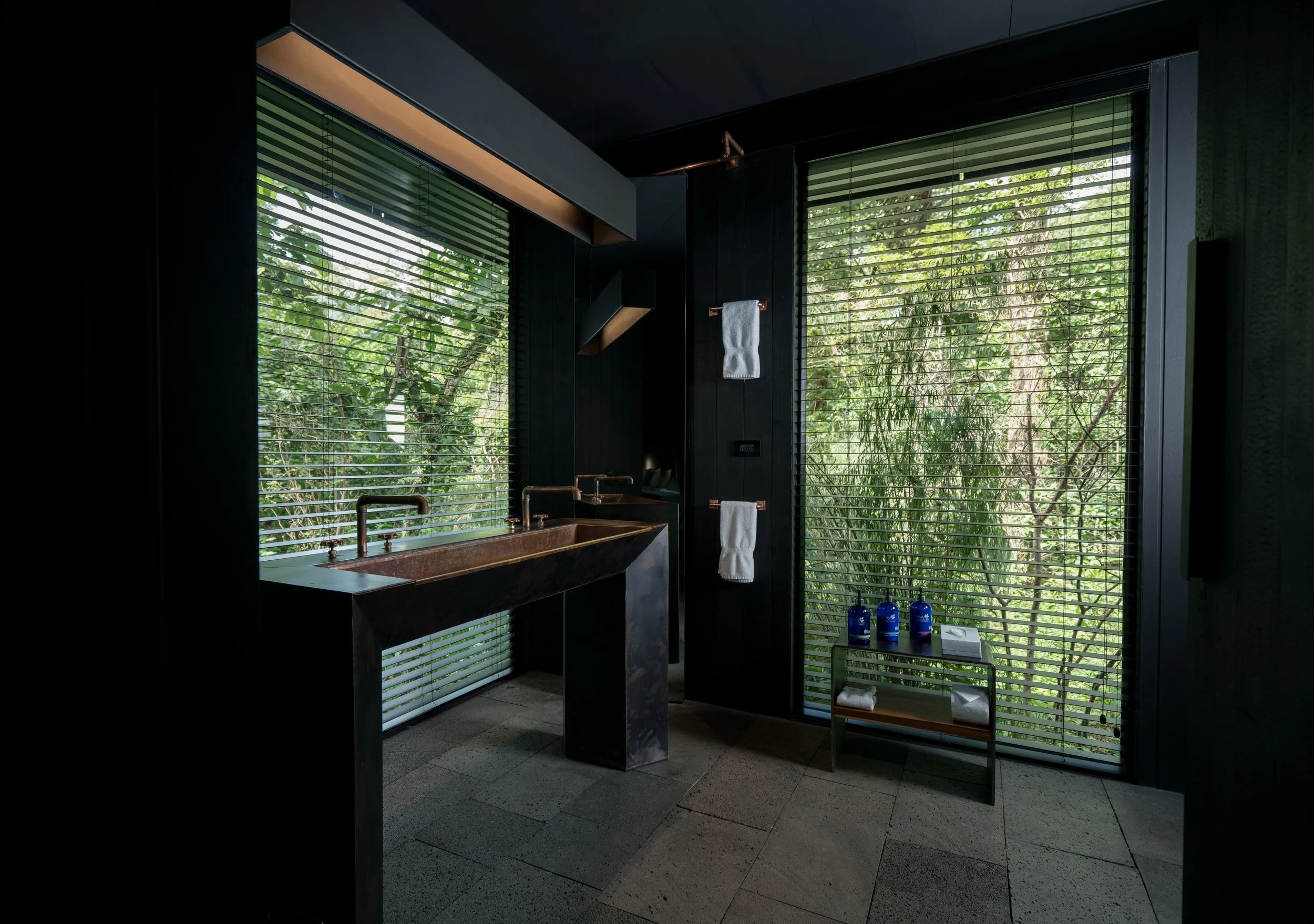 Modern bathroom with black walls and large windows with horizontal blinds, green view outside, copper sinks, and a small metal table with blue bottles and folded towels.