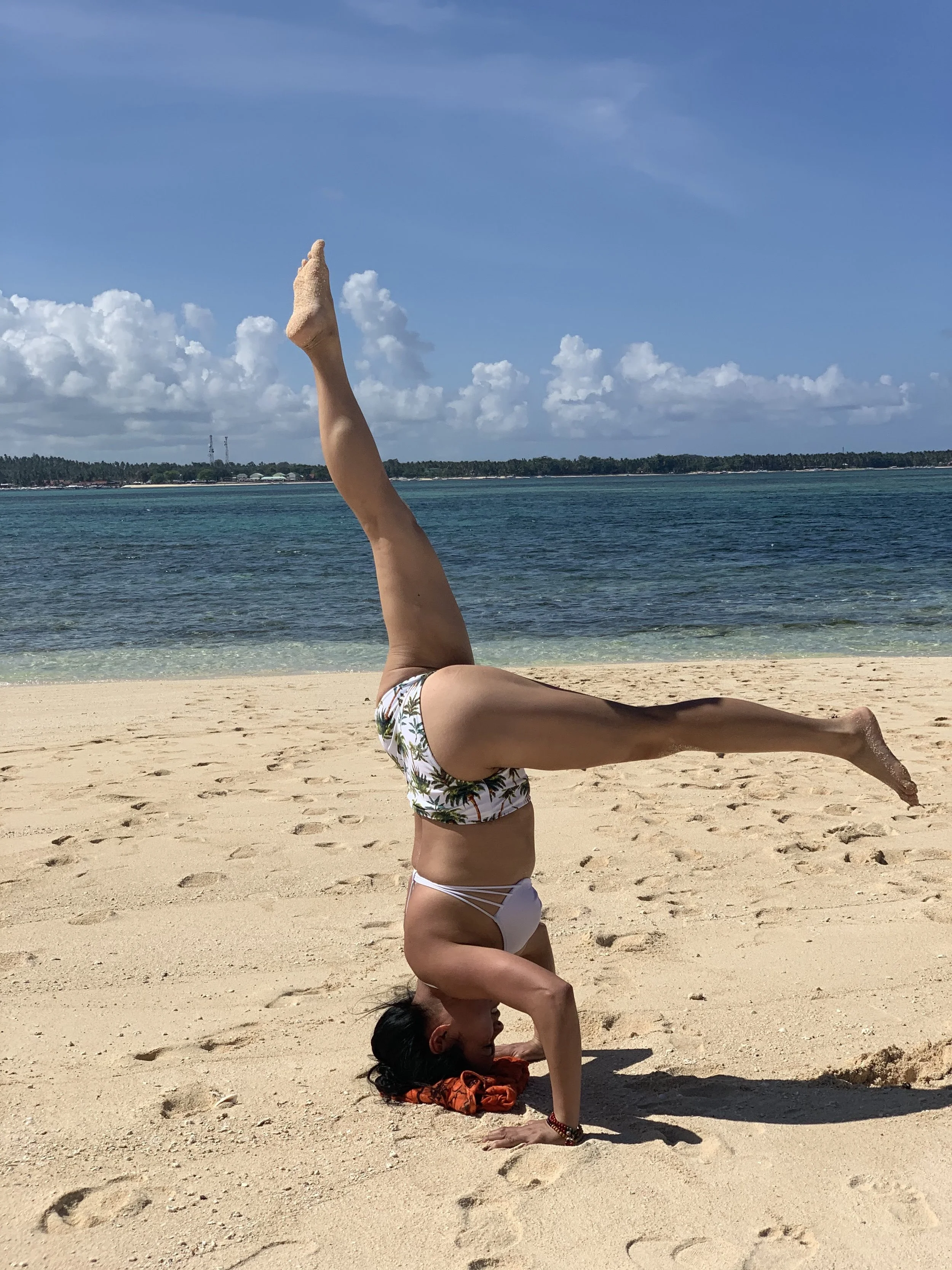 A woman practicing yoga or acrobatics on a sandy beach, performing a headstand with one leg raised upwards and the other extended outward, near the ocean with a cloudy sky in the background.