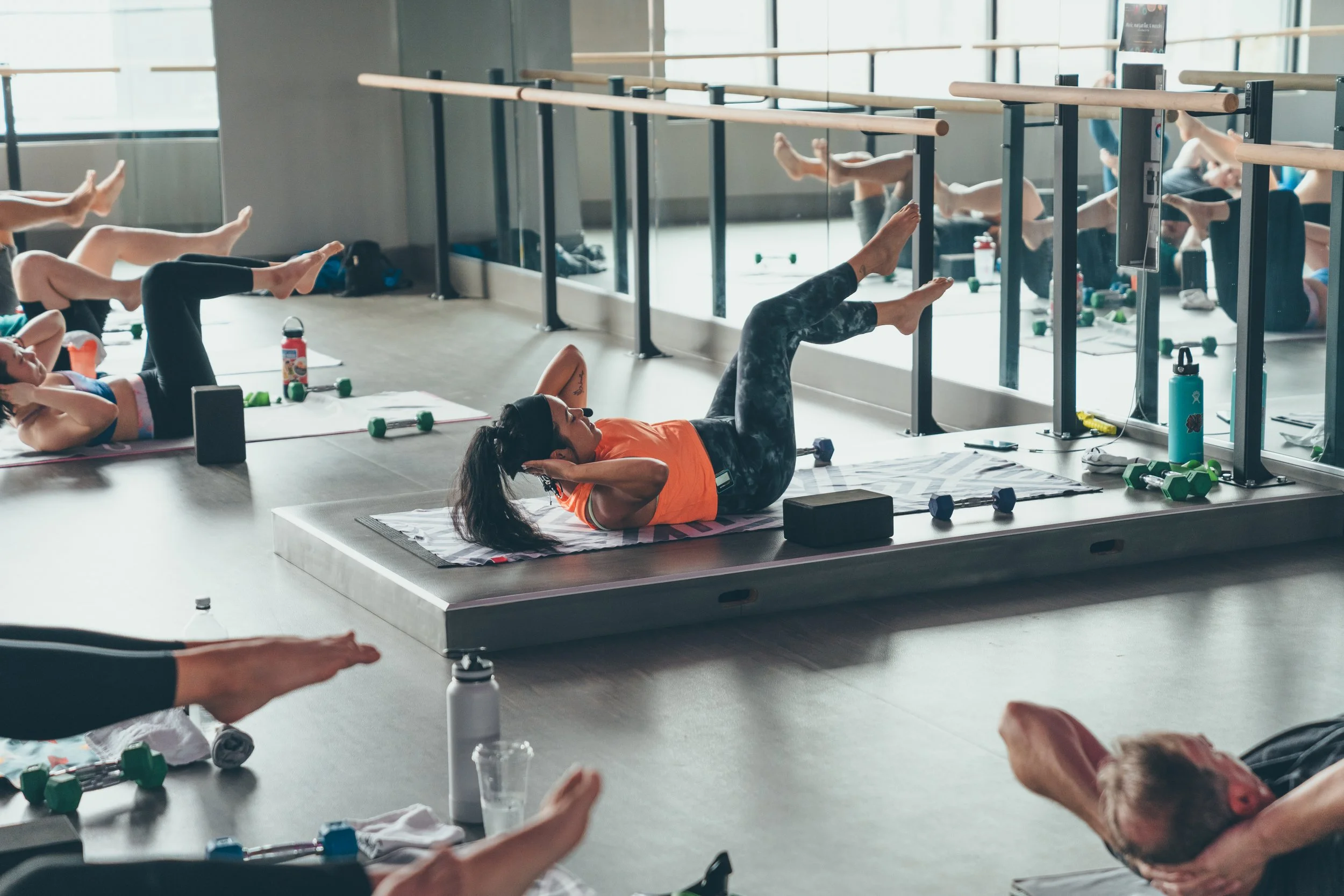 Group of people participating in a fitness or yoga class in a bright studio, lying on mats, stretching, and following instructor-led exercises with a woman in the foreground performing a core workout.