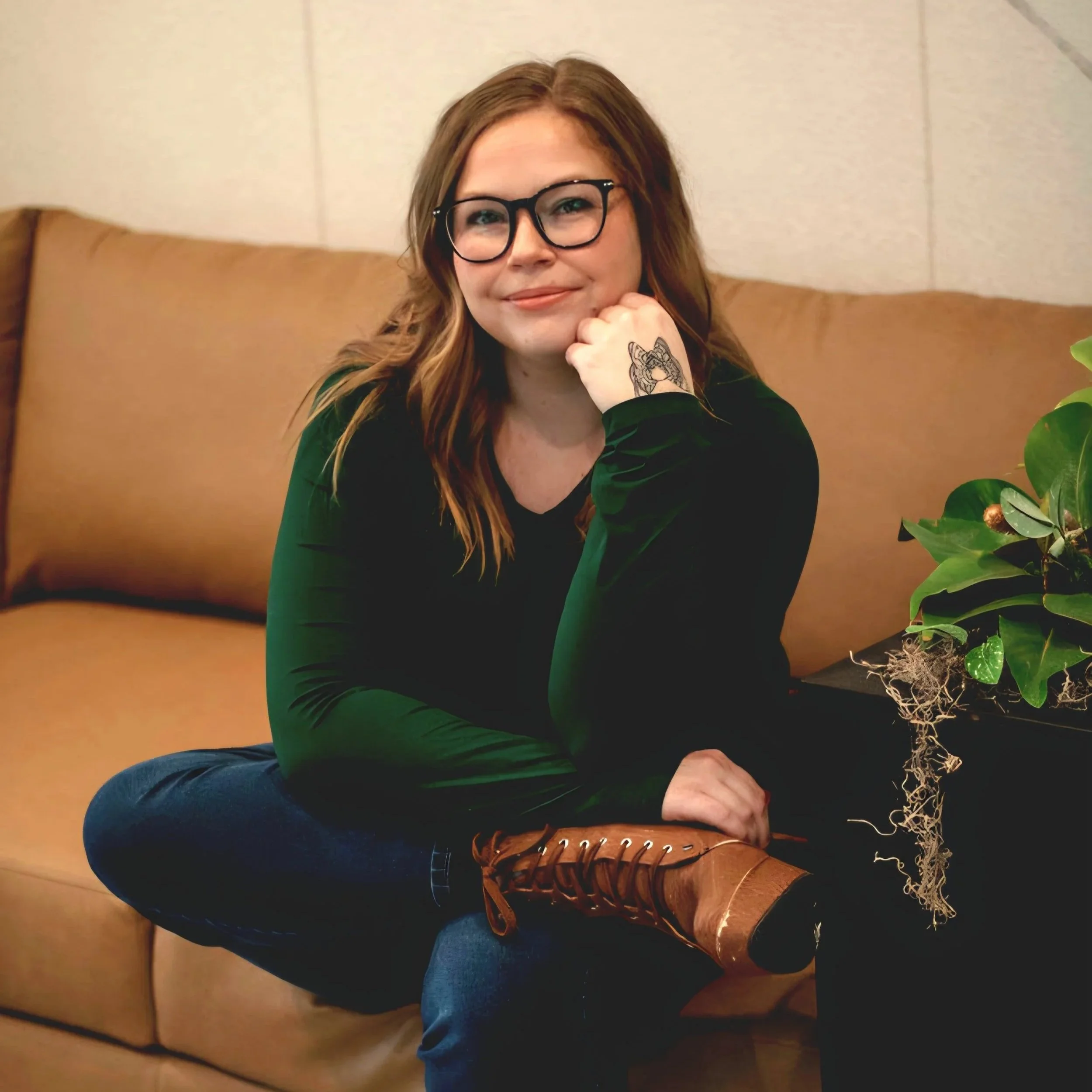 A woman with glasses and a tattoo on her wrist sitting on a tan couch, with a plant nearby.