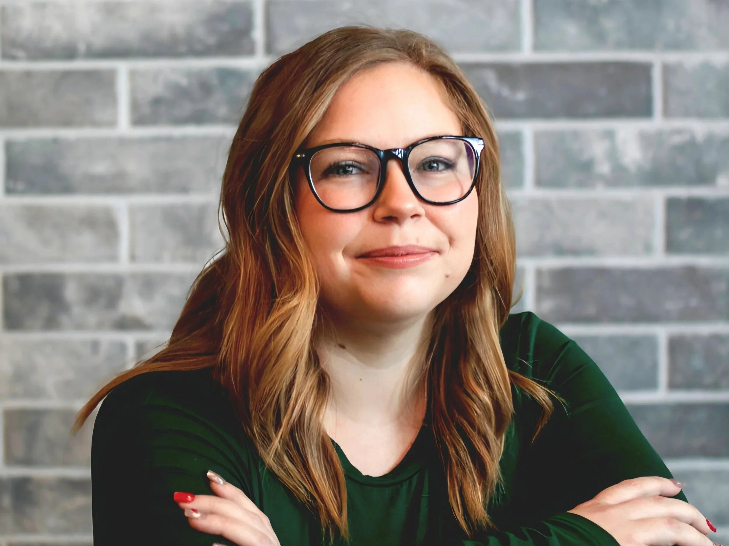 A woman with red hair, glasses, and a black top sitting in front of a gray brick wall, smiling with crossed arms.