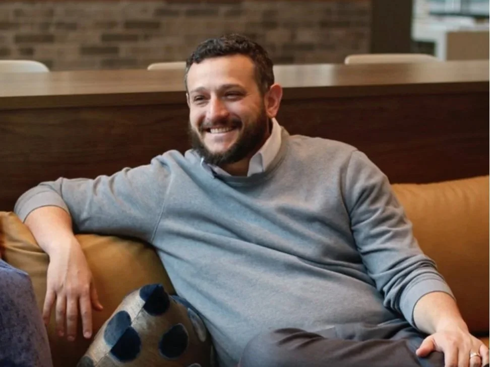 A man with a beard and short dark hair smiling and sitting on a tan cushion in a cozy indoor setting, with a brick wall in the background.