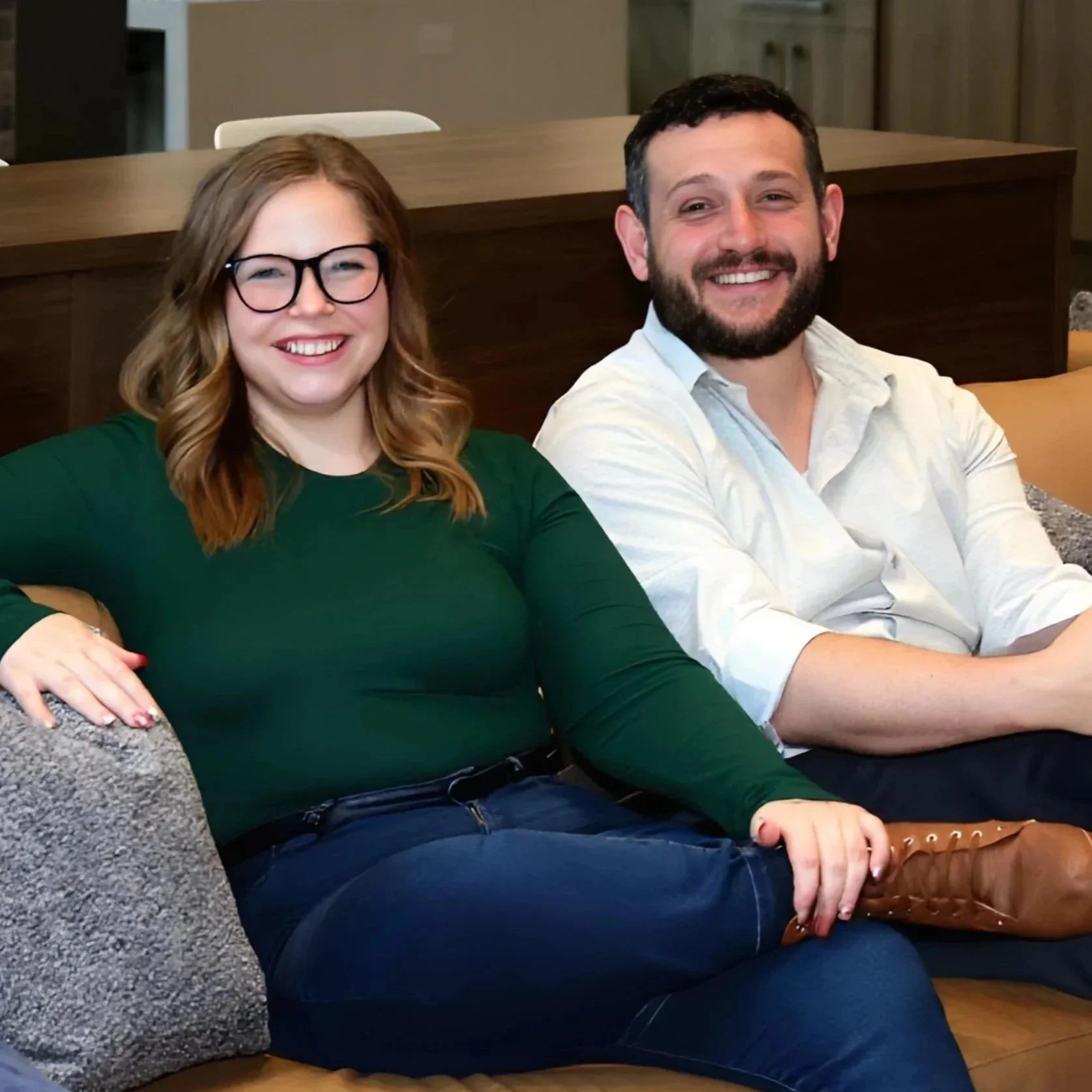 A smiling woman with glasses wearing a green top and jeans, sitting on a sofa with a man in a white shirt, both relaxed and happy.
