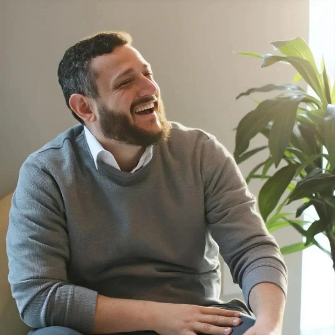 A man with a beard and short dark hair laughing while sitting next to a large green plant, wearing a gray sweater and white shirt, in a well-lit room.