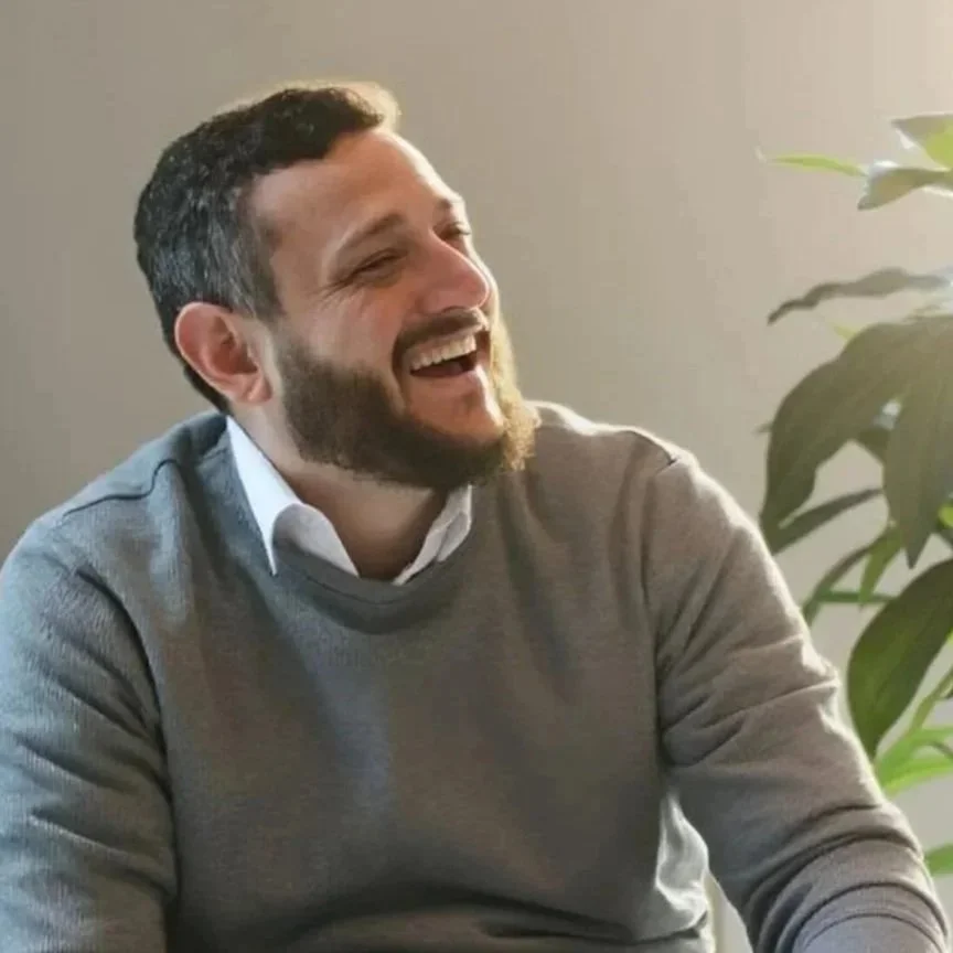 A man with dark hair and a beard is smiling and laughing while sitting at a table. He is wearing a gray sweater over a white collared shirt. There is a green plant on his right side and a plain wall in the background.