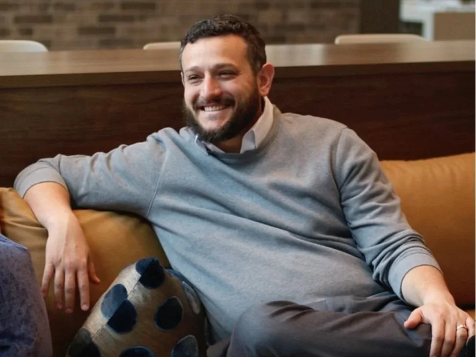 A man with a beard smiling and sitting relaxed on a beige couch, wearing a gray sweatshirt and a white collared shirt underneath, in a cozy indoor setting.