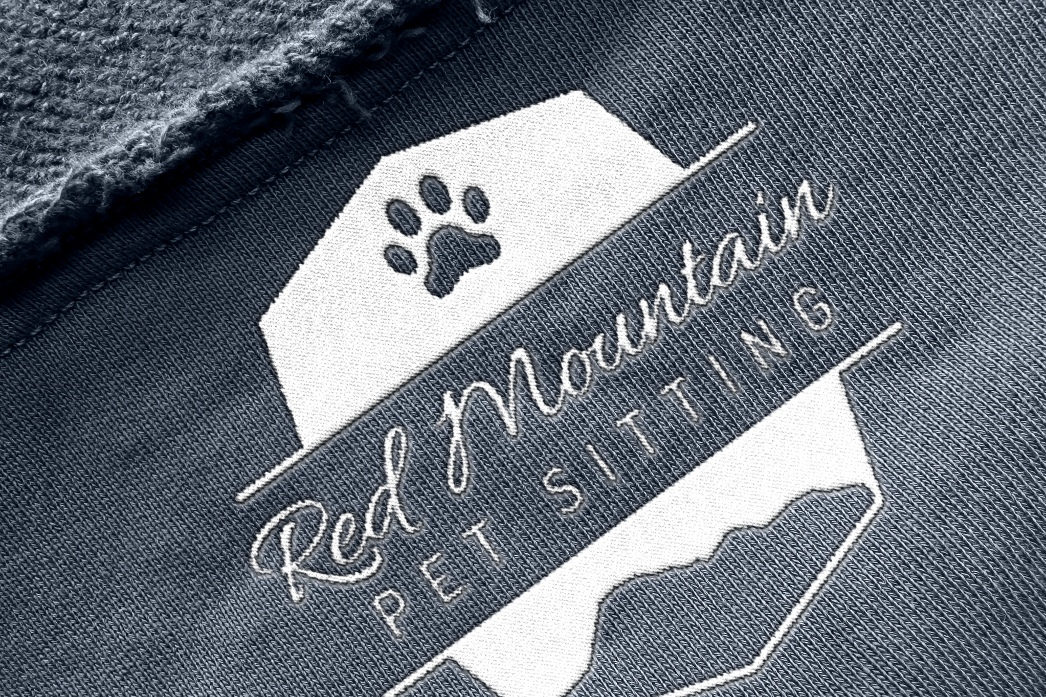 Close-up of an embroidered logo that features a dog paw print, a mountain silhouette, and the text 'Red Mountain Pet Sitting' on fabric.