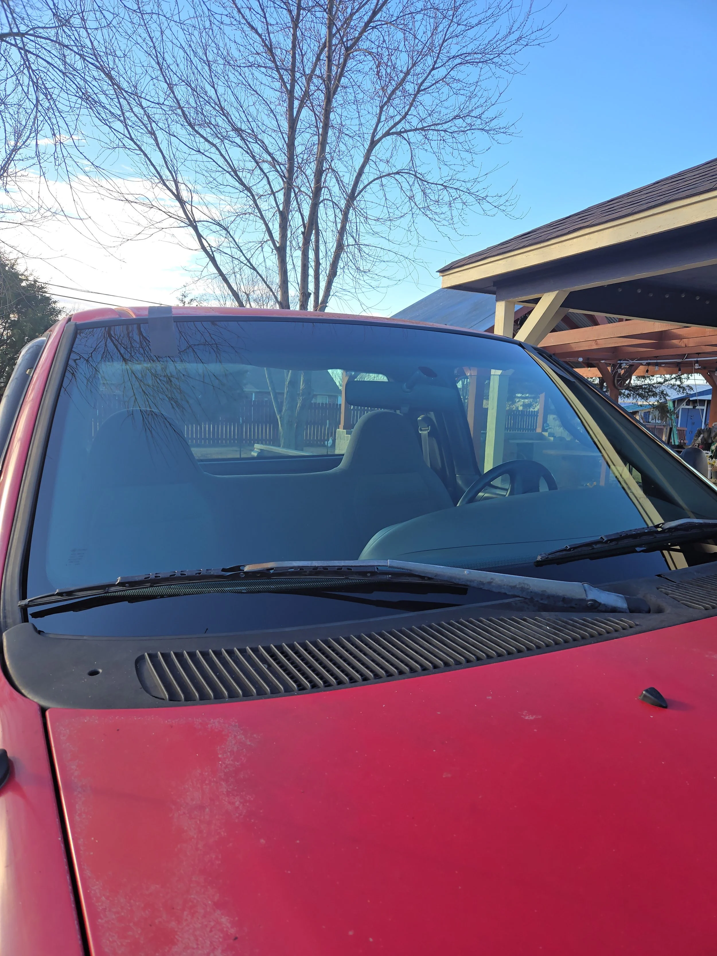 Red vintage vehicle with a cracked and dusty windshield, parked outdoors near a wooden structure, with leafless trees and a clear blue sky in the background.