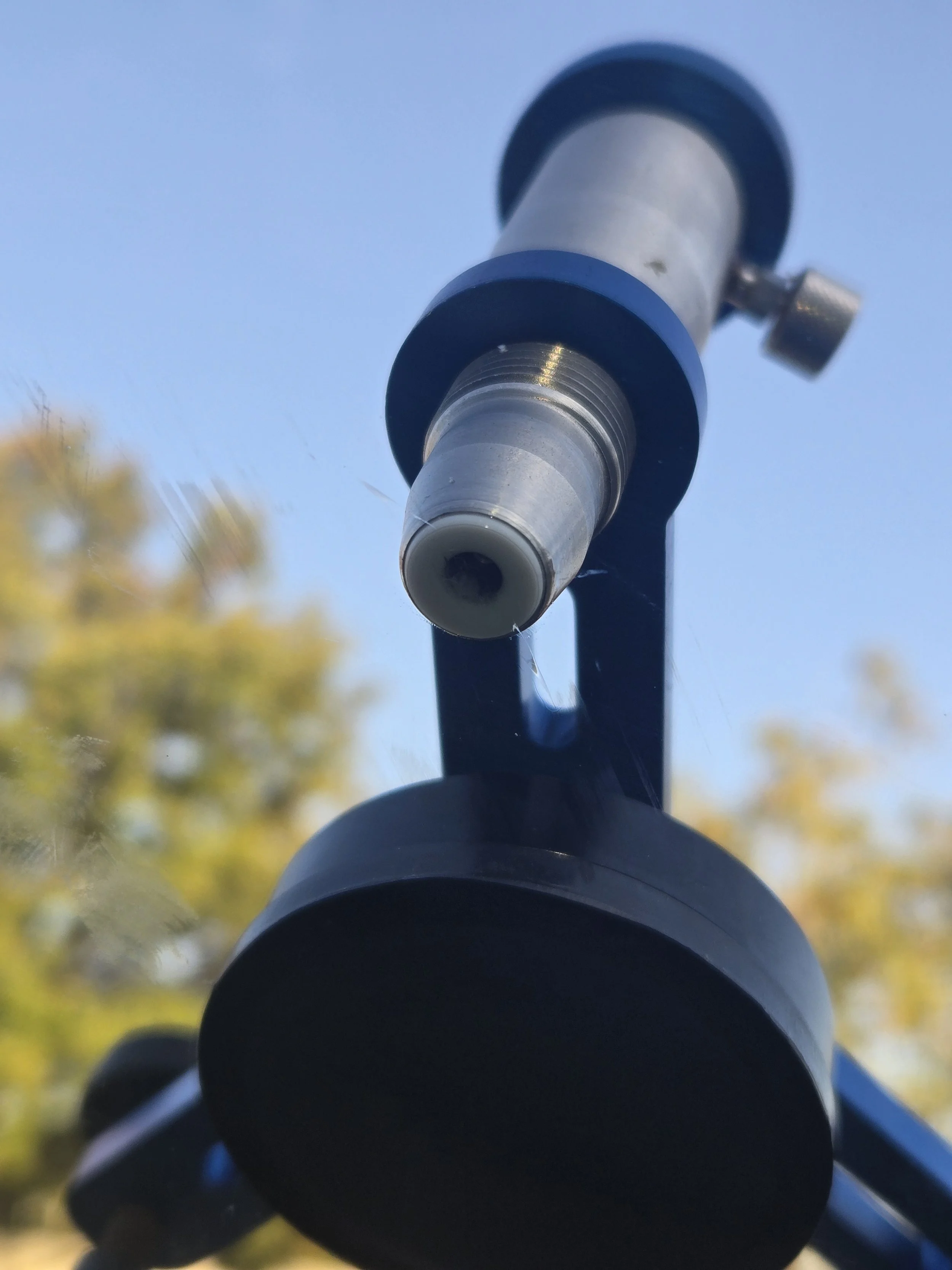 Close-up of an outdoor telescope mounted on a black stand, with a clear blue sky and blurred autumn trees in the background.
