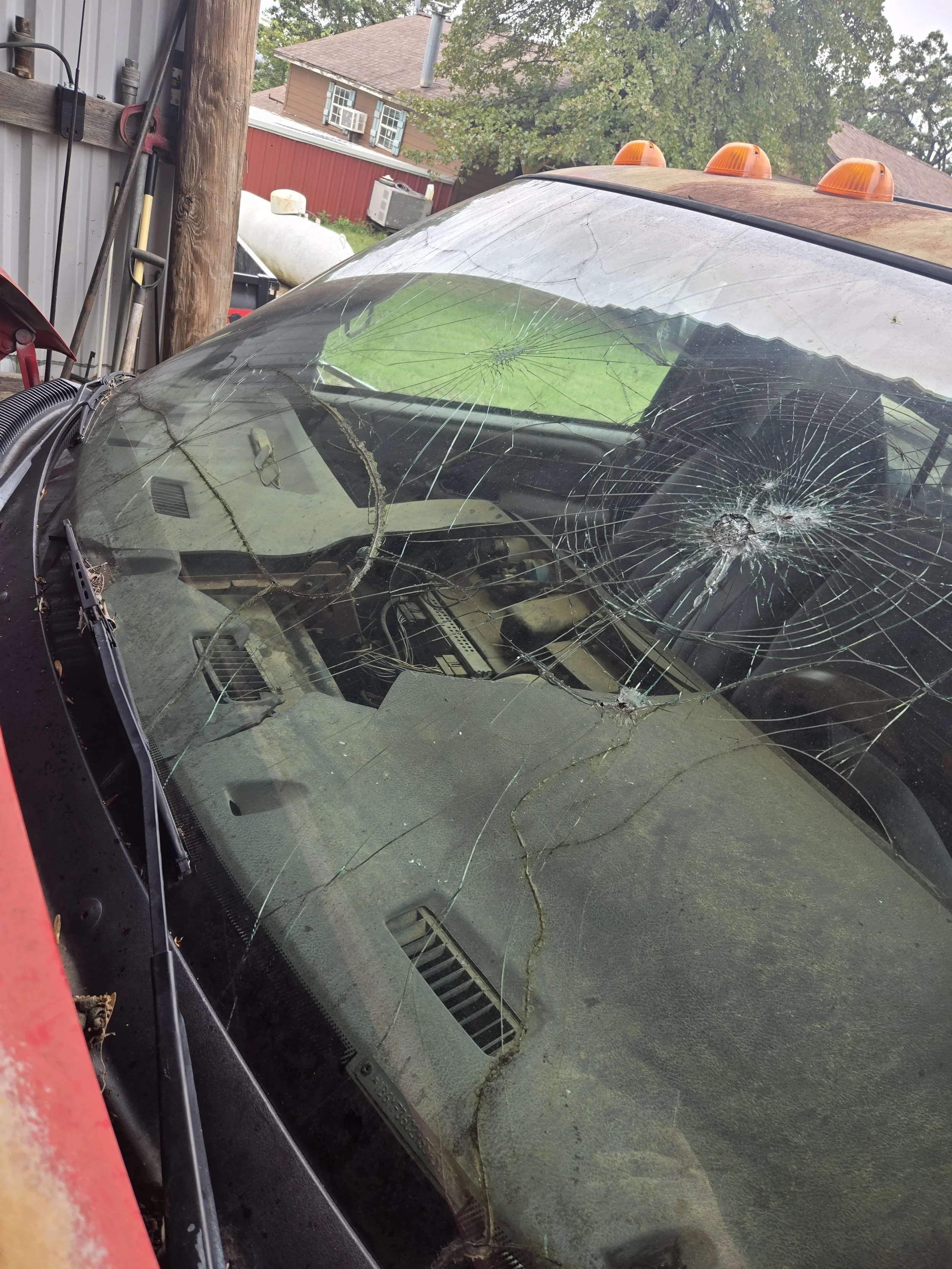 A car with a shattered windshield, multiple cracks and bullet hole-like damage, parked outdoors near a metal shed and wooden utility pole.