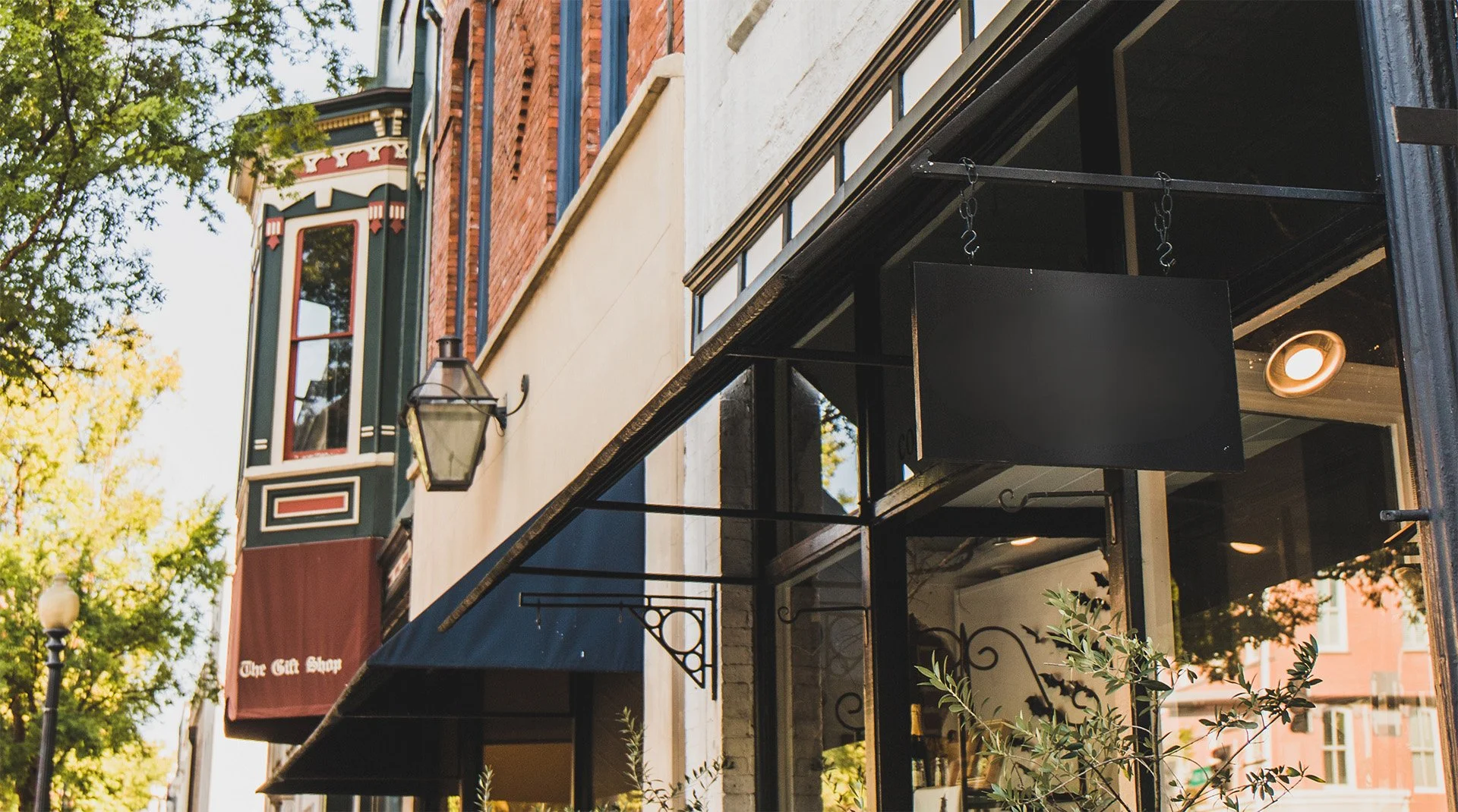 Street view of a storefront with black awnings and a hanging lantern in a tree-lined neighborhood