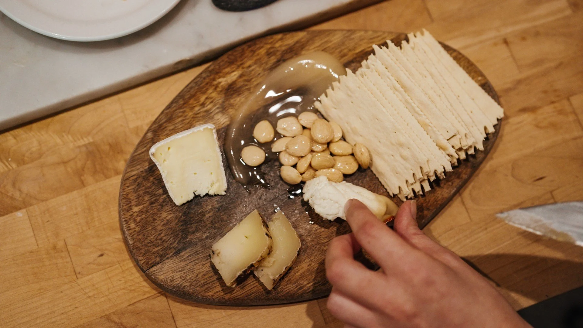A wooden serving board with wedges of cheese, macadamia nuts, and flat crispy crackers