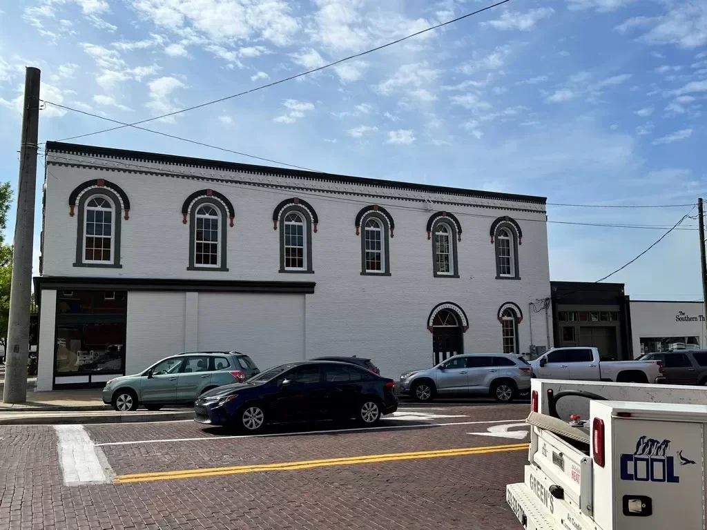 White historic building with arched windows, decorative trim, and cars parked in front