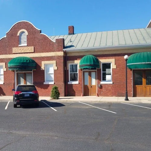 Historic brick train depot building with green awnings, a "BAGGAGE" sign, and a parked SUV