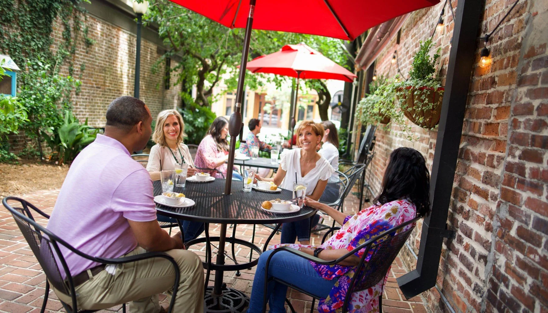 People dining at outdoor restaurant patio with red umbrellas, enjoying meals and conversation