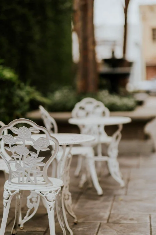 White ornate metal patio chairs and tables on a wooden deck, with a blurred background