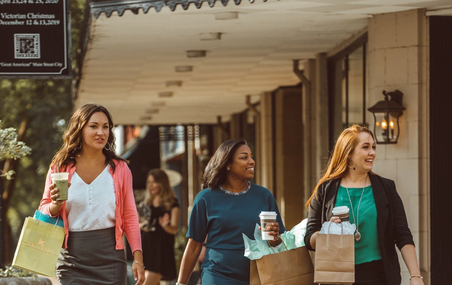 Three women walking along a city sidewalk with shopping bags and drinks, smiling and talking