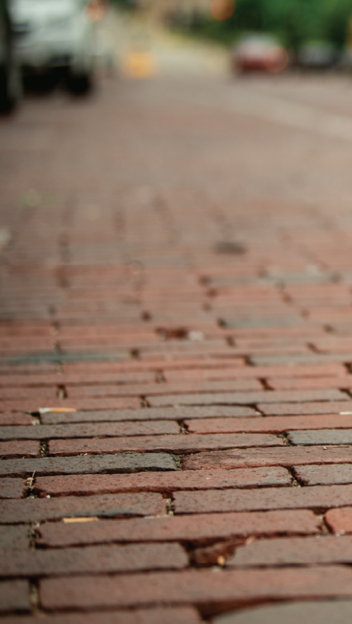 Close-up view of a brick sidewalk with some cars parked in the background, slightly out of focus.