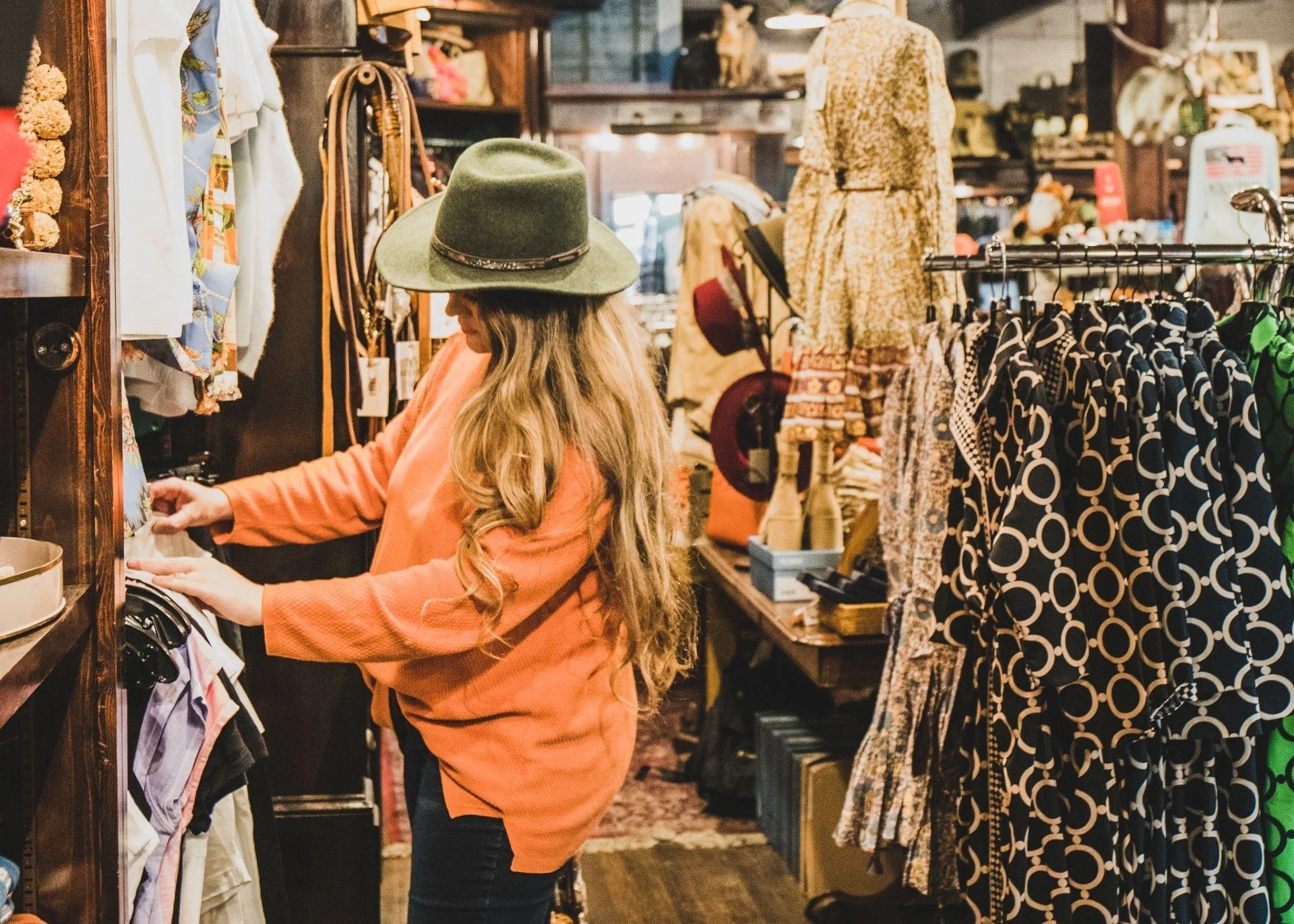 A woman shopping in a boutique clothing store, wearing a wide-brimmed green hat and an orange jacket