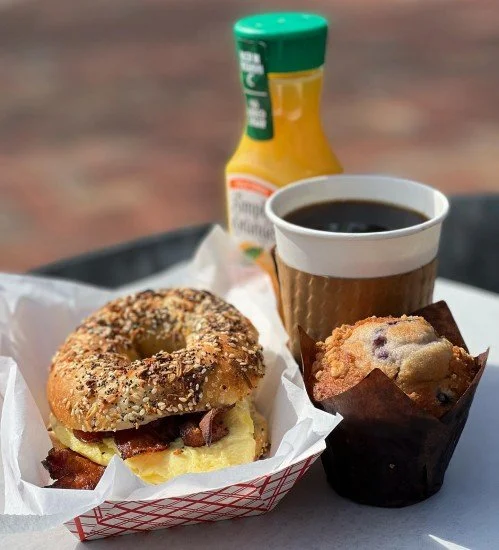Paper basket with a bacon, egg, and cheese breakfast sandwich next to a muffin and black coffee