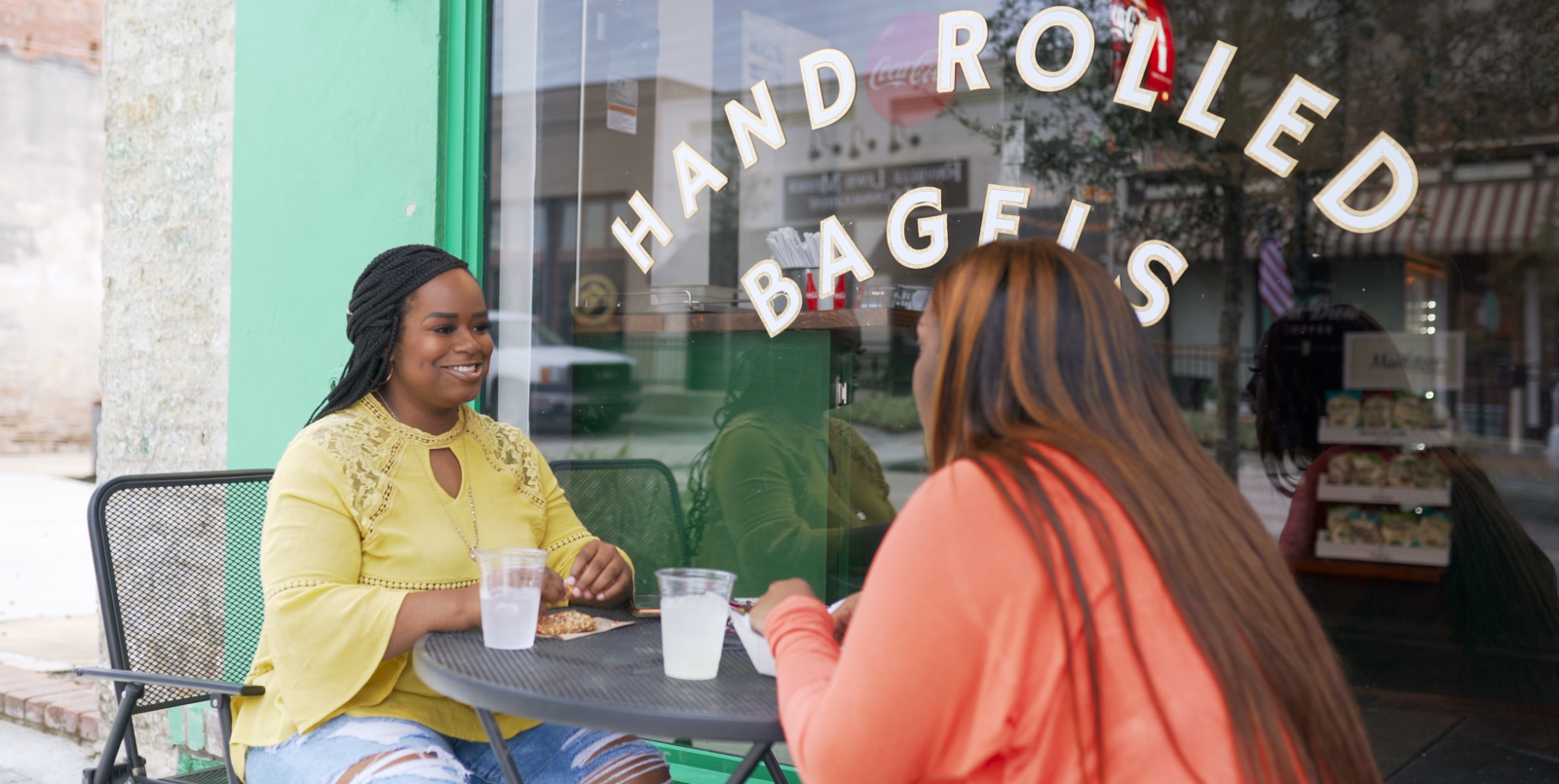 Two women sitting outside a restaurant called 'Hand Rolled Bagels'