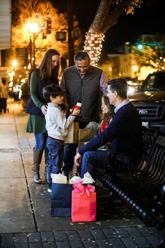 Family at night on city sidewalk near a decorated tree, exchanging Christmas gifts