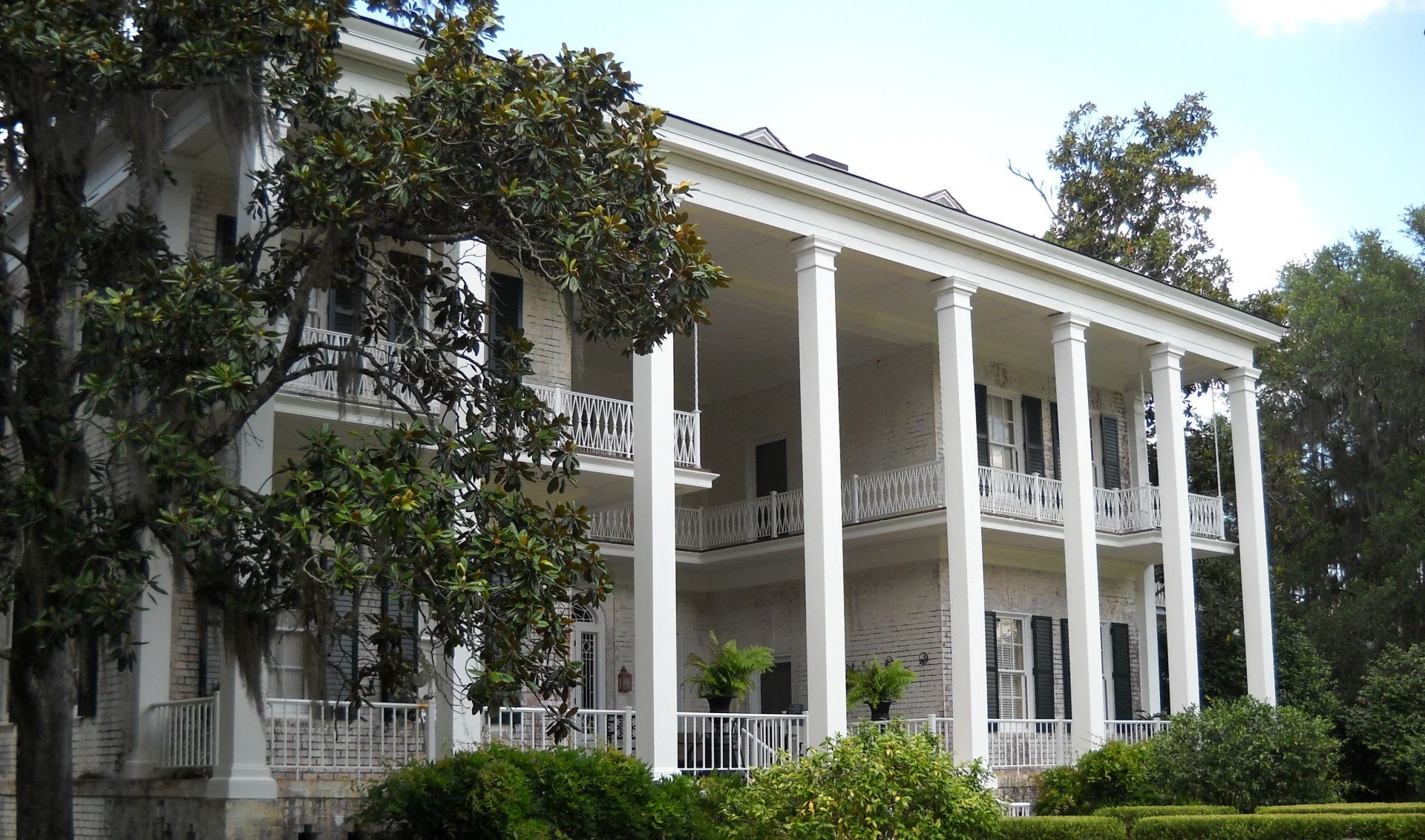 Large white house with tall columns supporting a front porch, surrounded by green trees and bushes