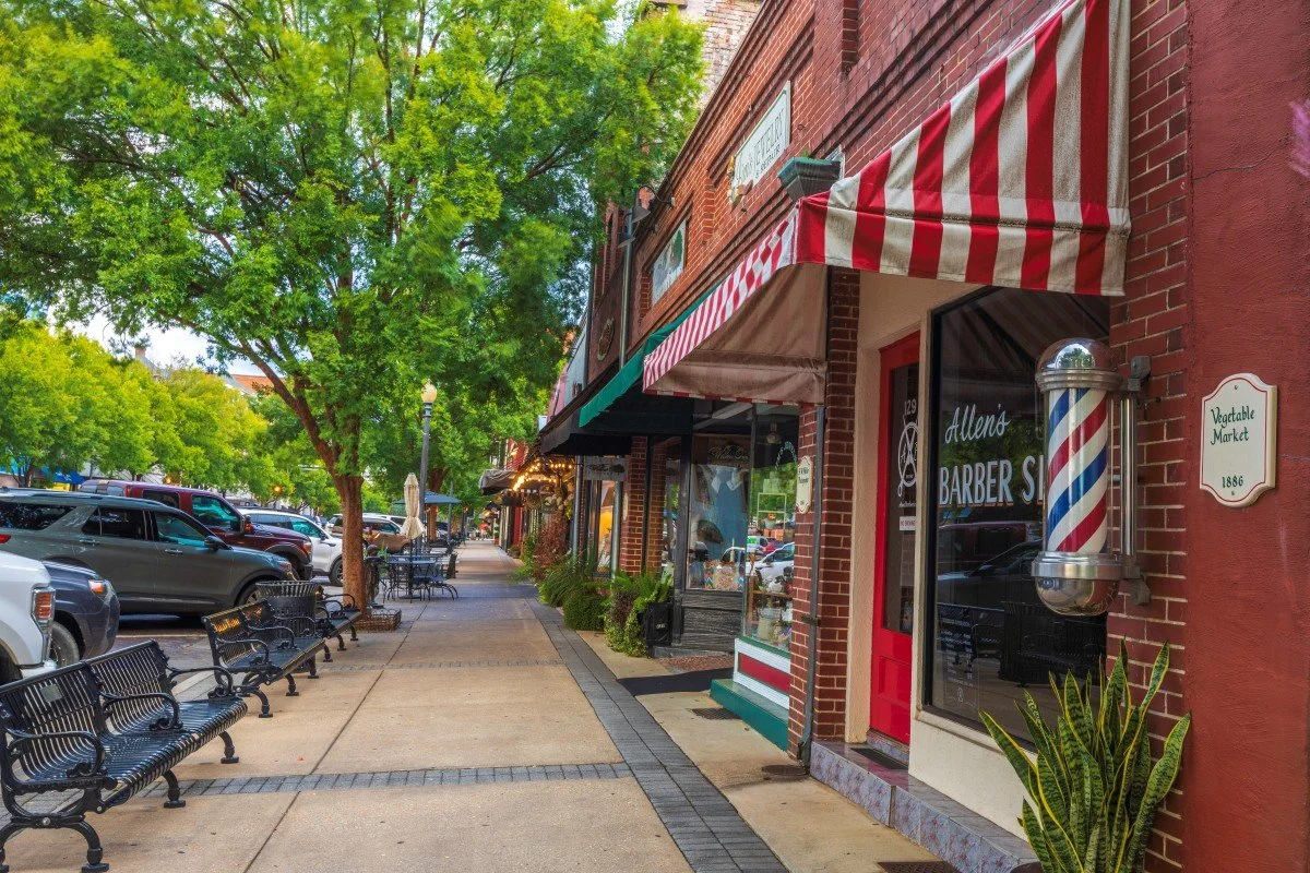 Downtown sidewalk with parked cars, green trees, and storefronts with green and red awnings