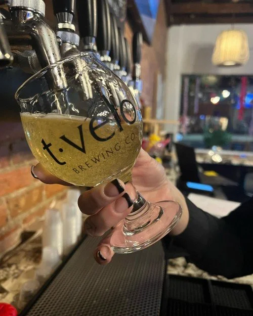 A hand holding up a glass beer chalice printed with a stylized "t-velo" logo