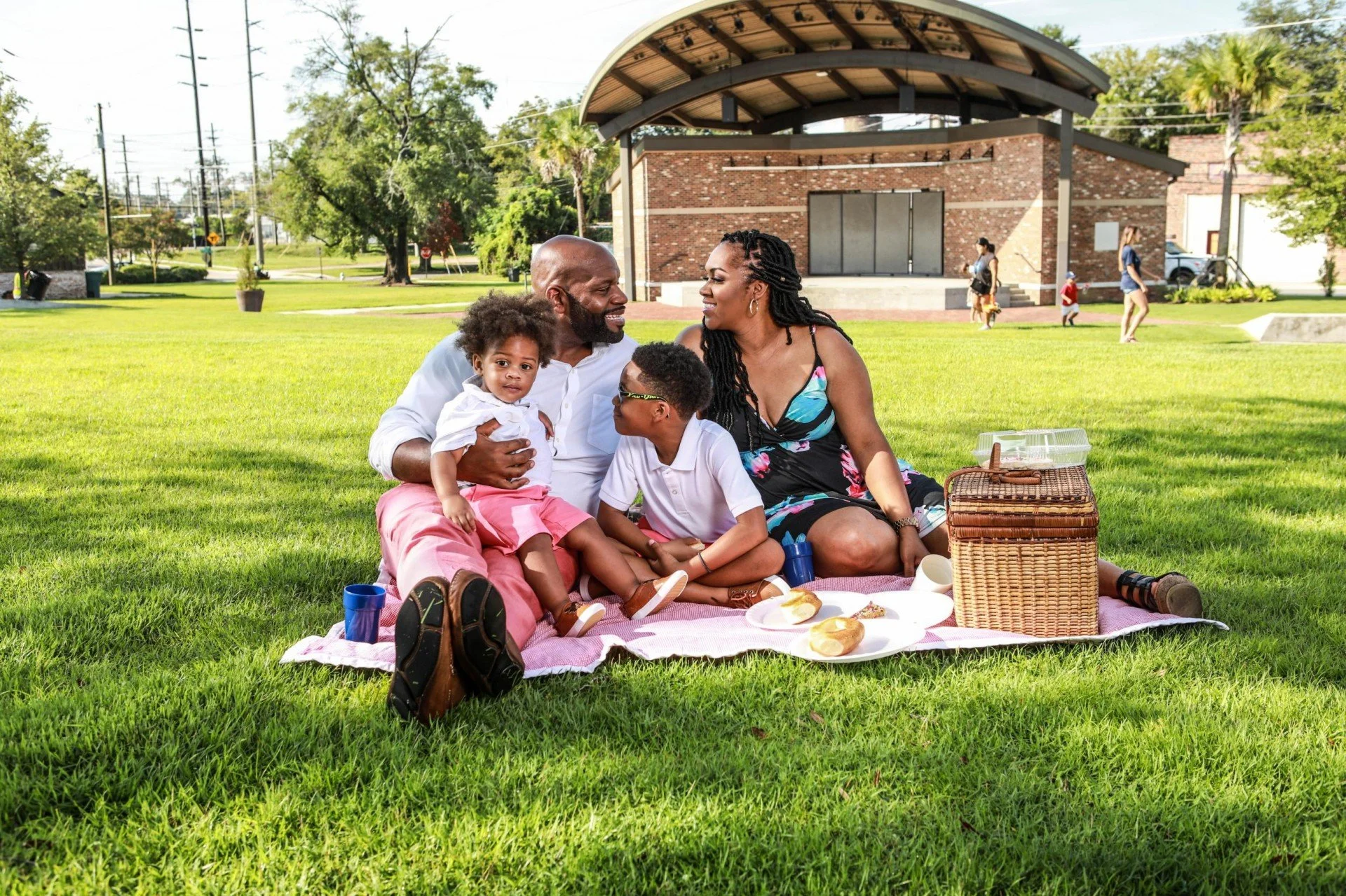 A family of four sitting on a blanket in a park enjoying a picnic outside of a pavilion
