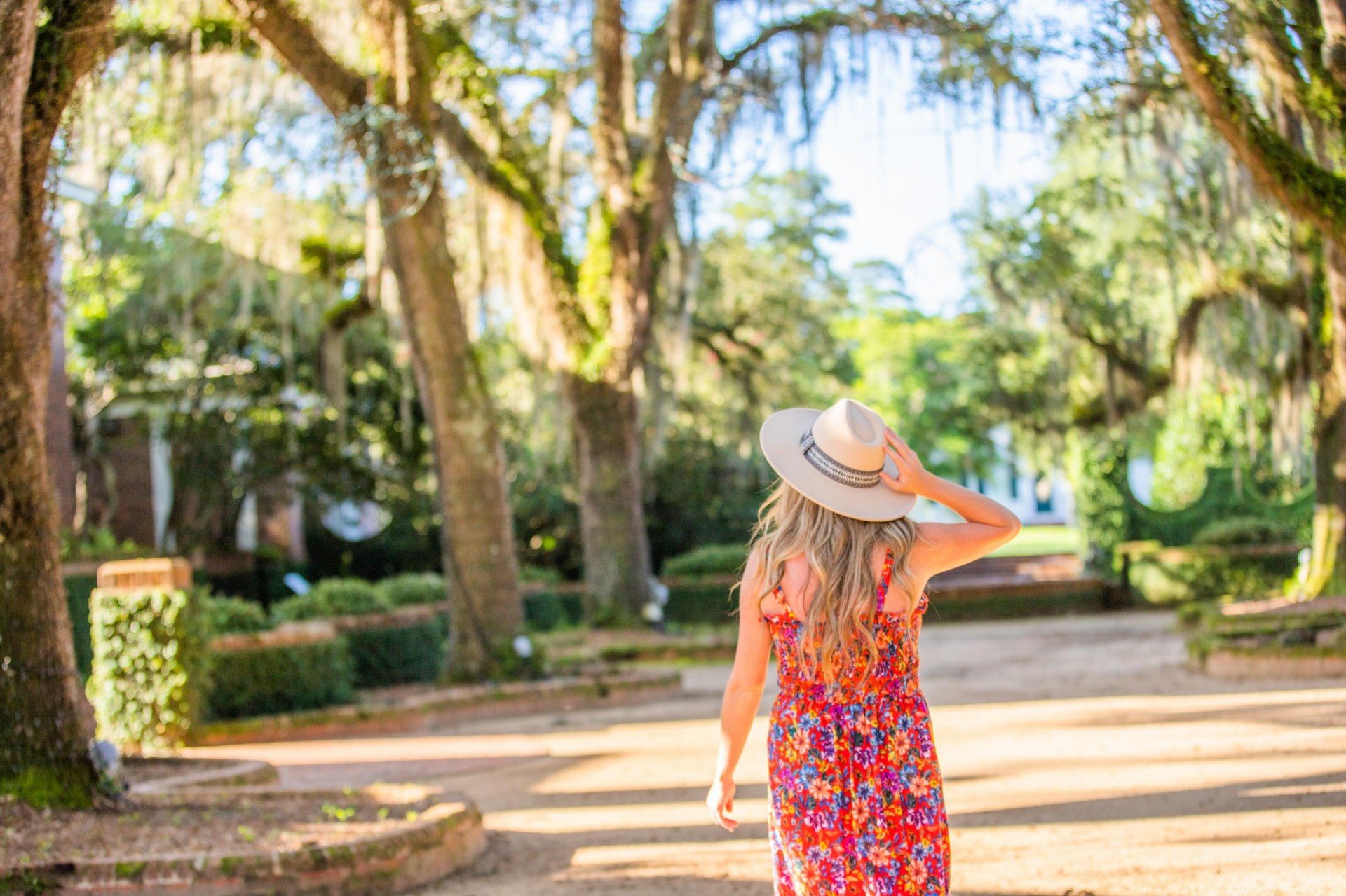 A woman with long wavy hair wearing a red floral dress and a wide-brimmed hat walking in a park