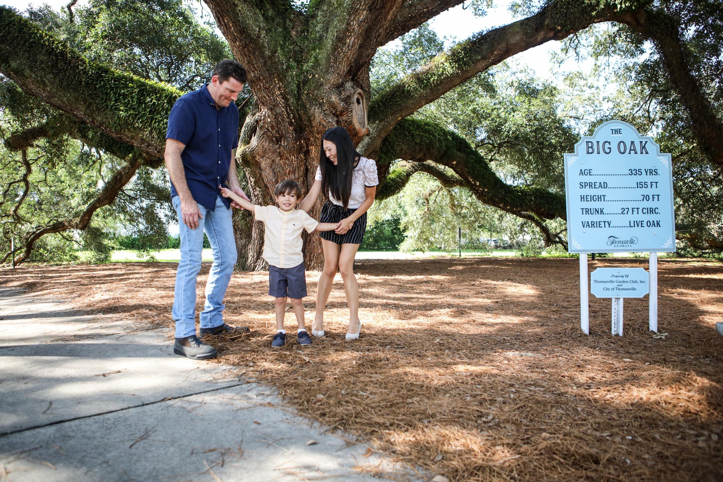 A family of three standing outdoors under a large oak tree, holding hands and smiling