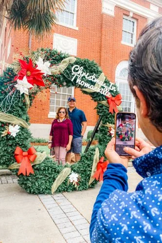 A person taking a photograph of a couple standing behind a large decorated Christmas wreath