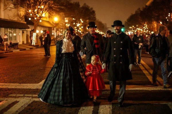 A family dressed in Victorian-style clothing walking on a lit-up street at night
