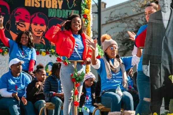 A group of smiling people riding on a Black History Month parade float, waving and celebrating