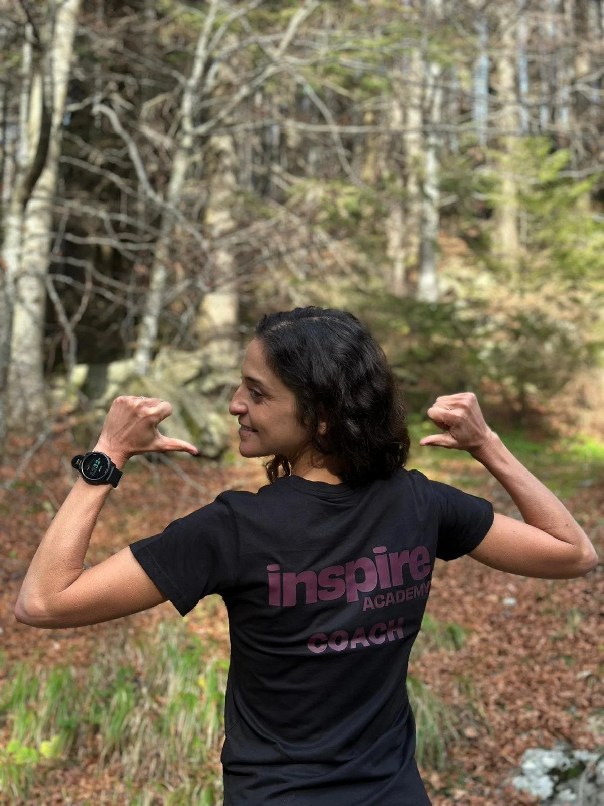 Donna che mostra i muscoli con un sorriso in un bosco, indossa una maglietta nera con la scritta 'inspire ACADEMY COACH' e un orologio sportivo