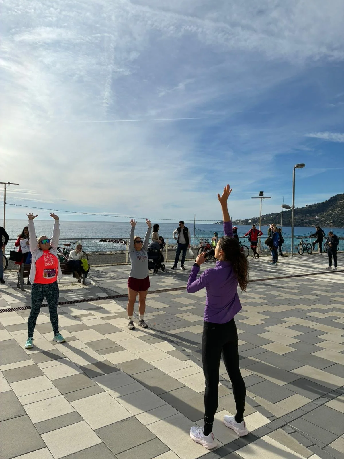 Gruppo di persone che fanno esercizi di stretching sulla passeggiata vicino al mare di giorno, con cielo sereno e alcuni ciclisti in lontananza.