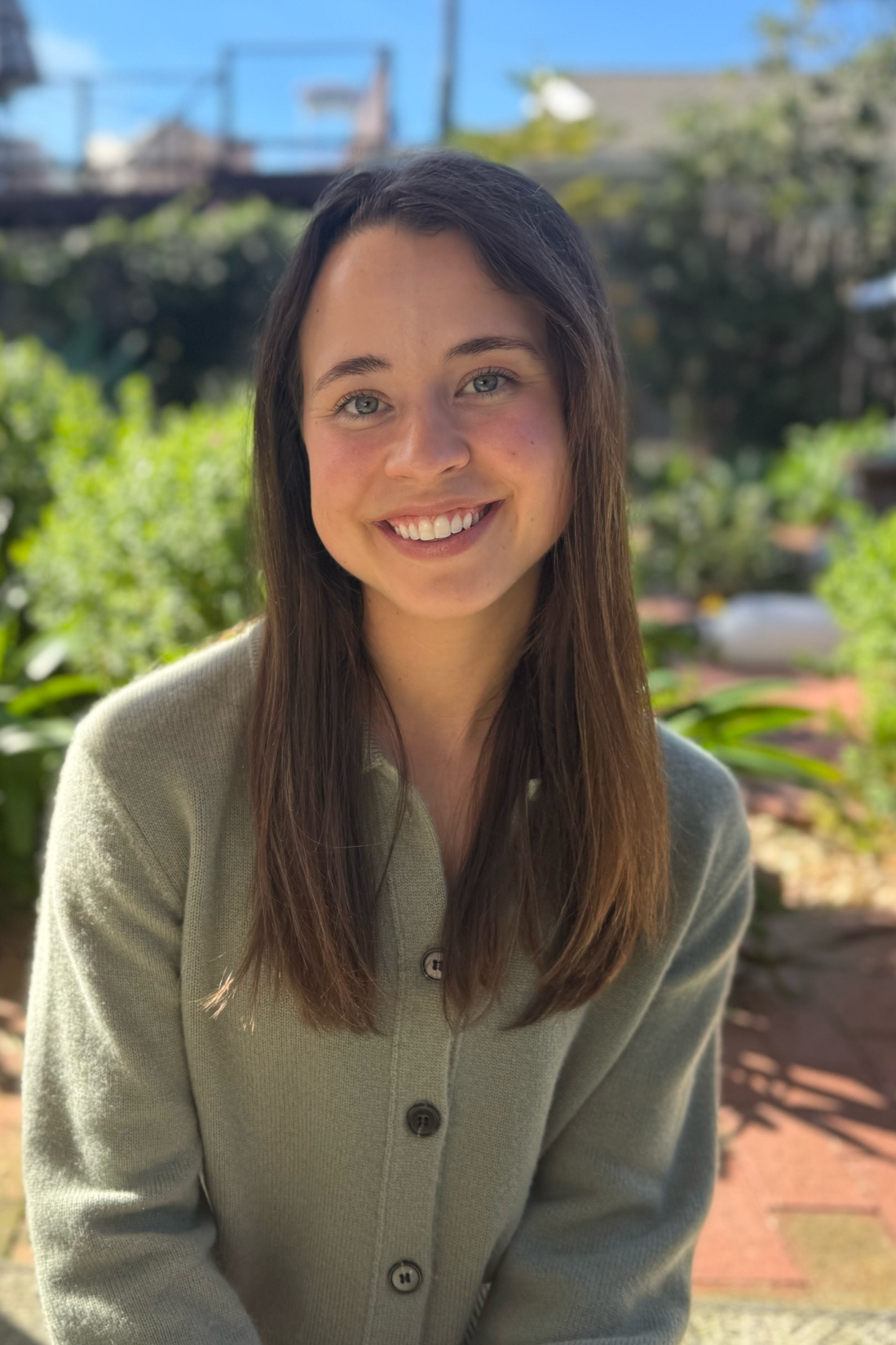 A young woman with long brown hair and blue eyes smiling outdoors on a sunny day, wearing a light green cardigan, with greenery and a blue sky in the background.