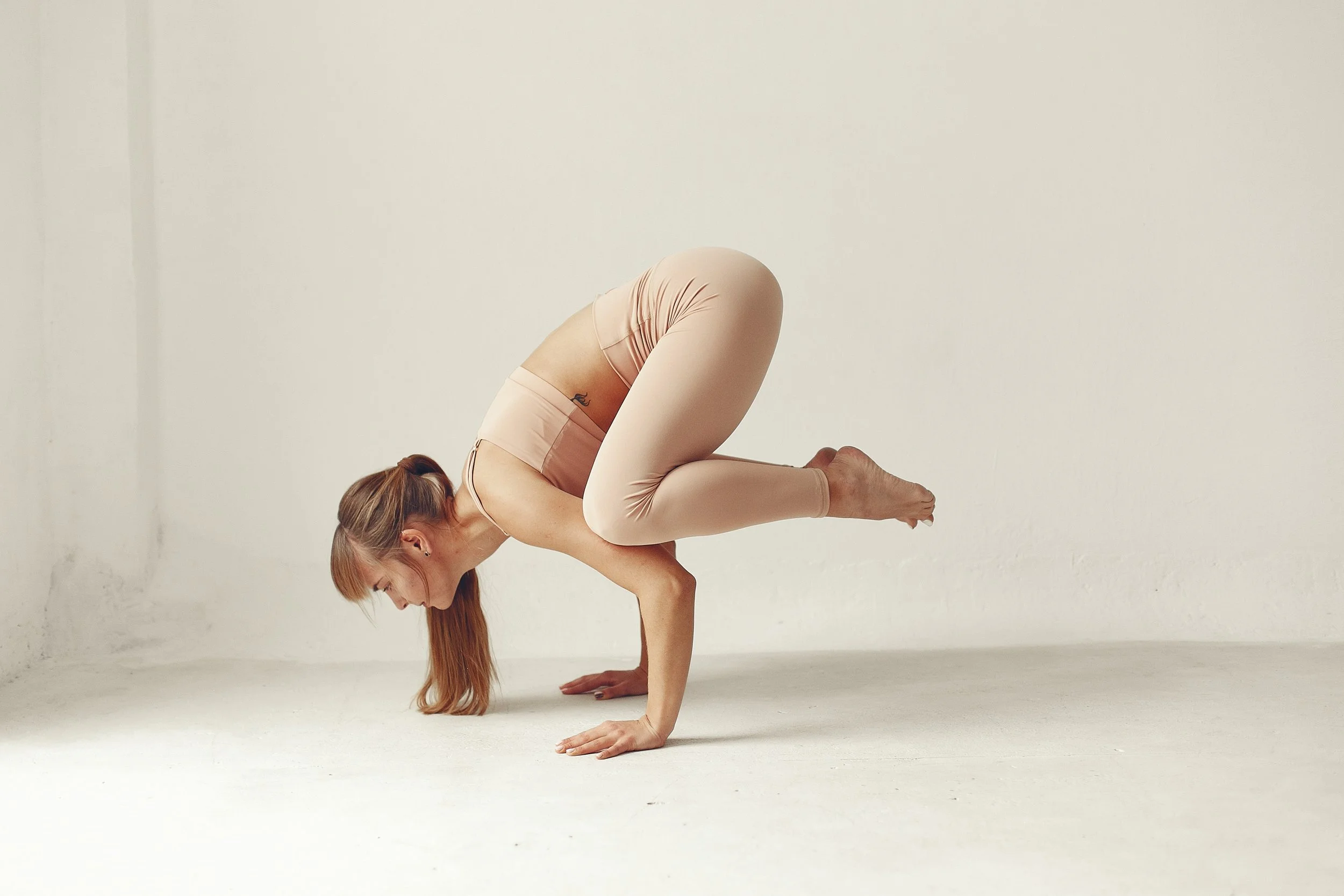 A woman in beige athletic clothing performing a handstand yoga pose in a minimalist white room.