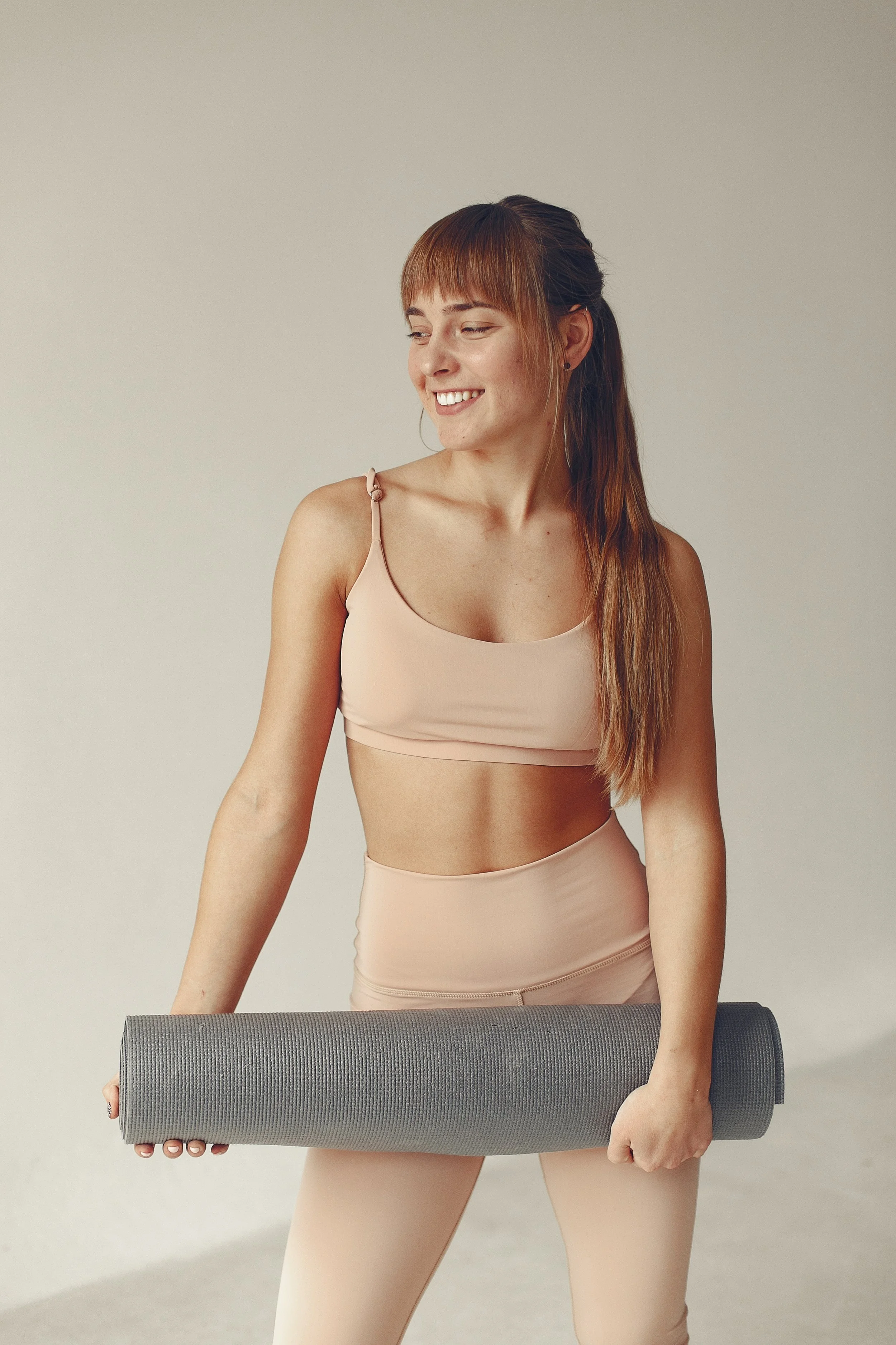 A woman in workout attire holding a rolled-up yoga mat, smiling and looking to her side.